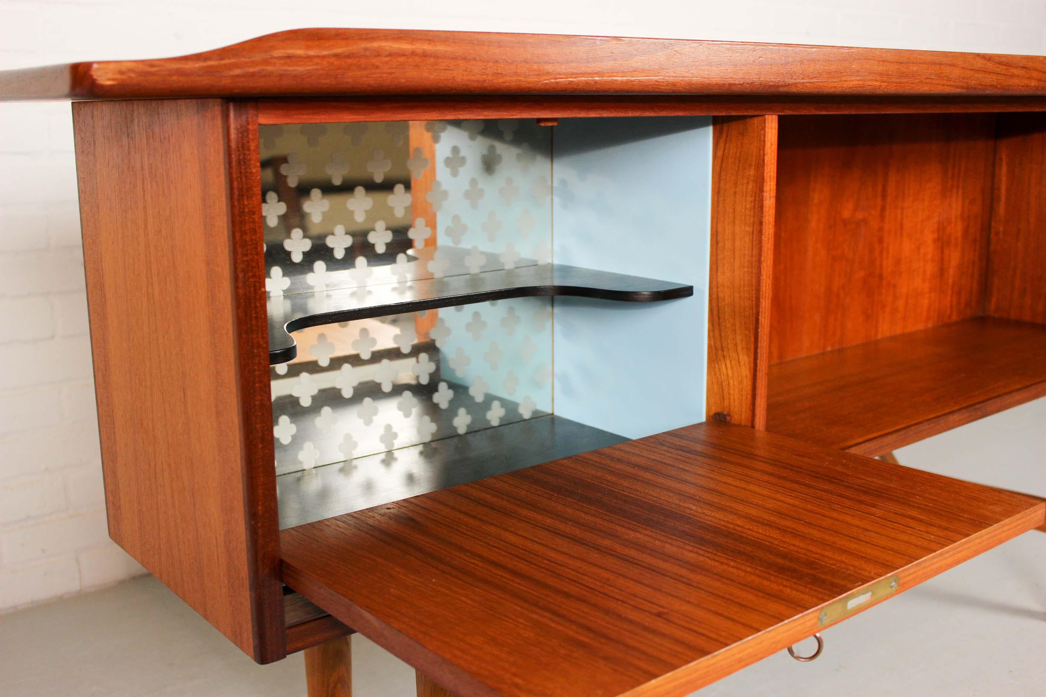 The desk is veneered in teak wood with solid teak legs and details. The frame has some brass parts. With beautiful drinks cabinet in light blue and bookshelve on backside. 
 