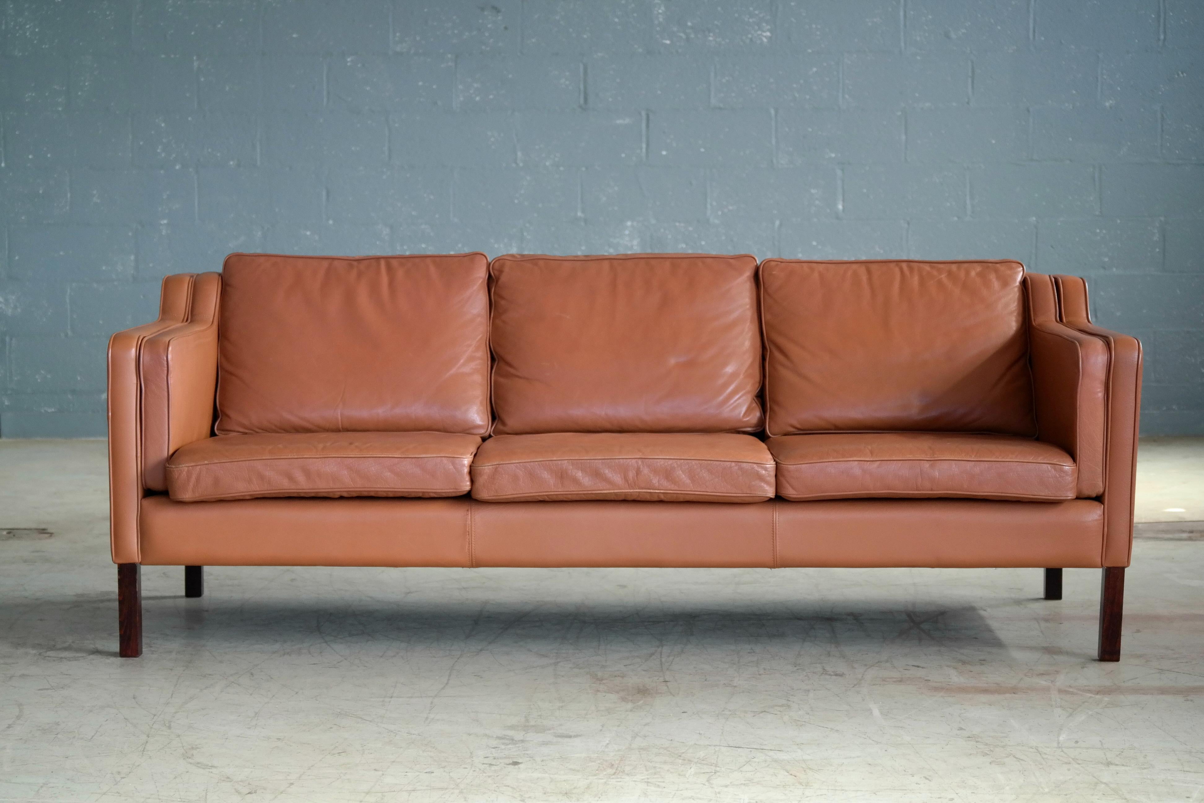 Classic Borge Mogensen style three-seat sofa in cognac leather by Stouby Mobler, Denmark. Buffalo hide raised on legs of beech. High quality craftsmanship and overall very good condition showing some natural age wear and patina with a couple of