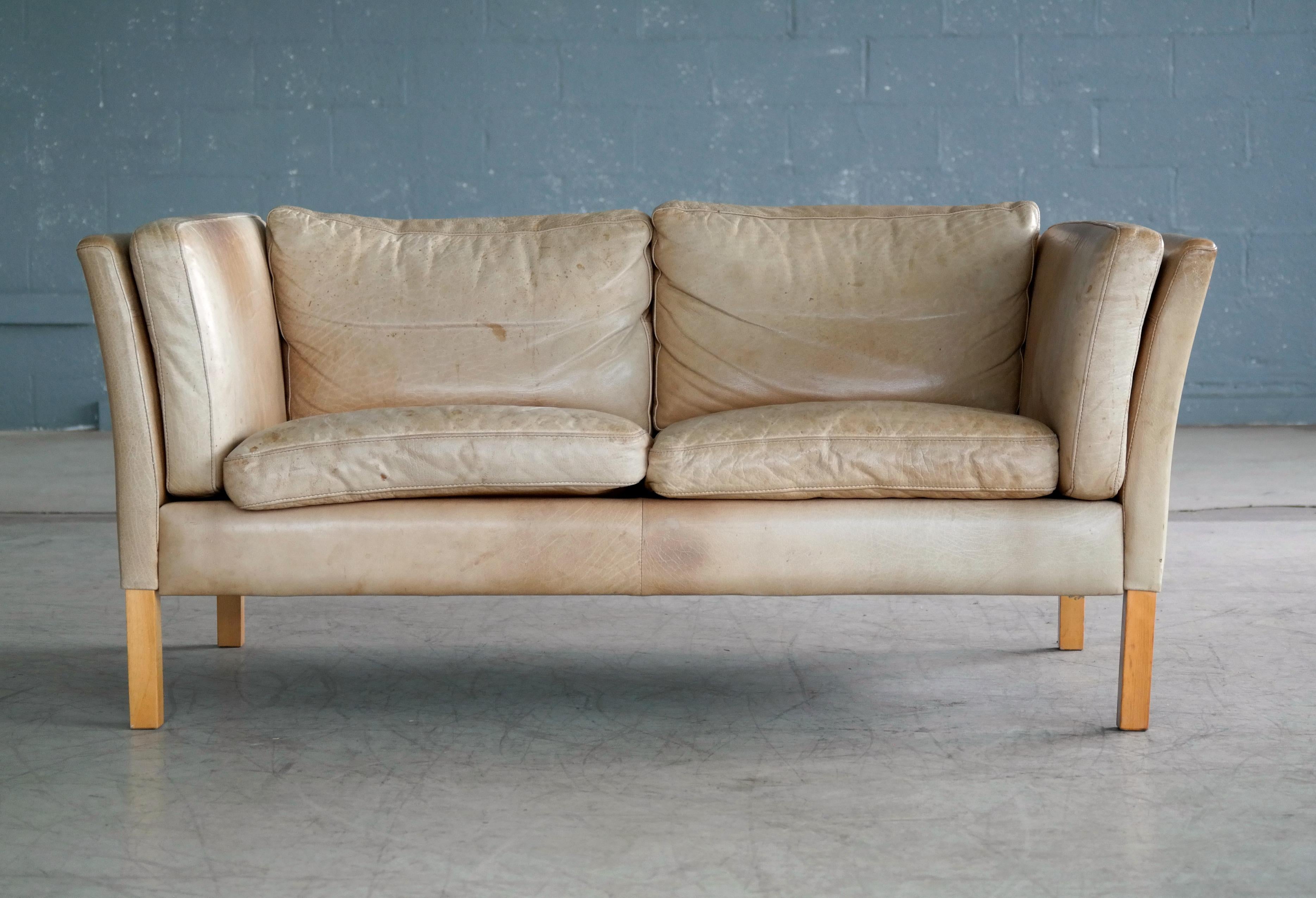 Great loveseat or two-seat sofa made by Stouby, Denmark. High quality construction blond beech wood frame around the armrests covered in a nice tan leather that has faded into an almost creme color over the decades. Significant patina and noble wear