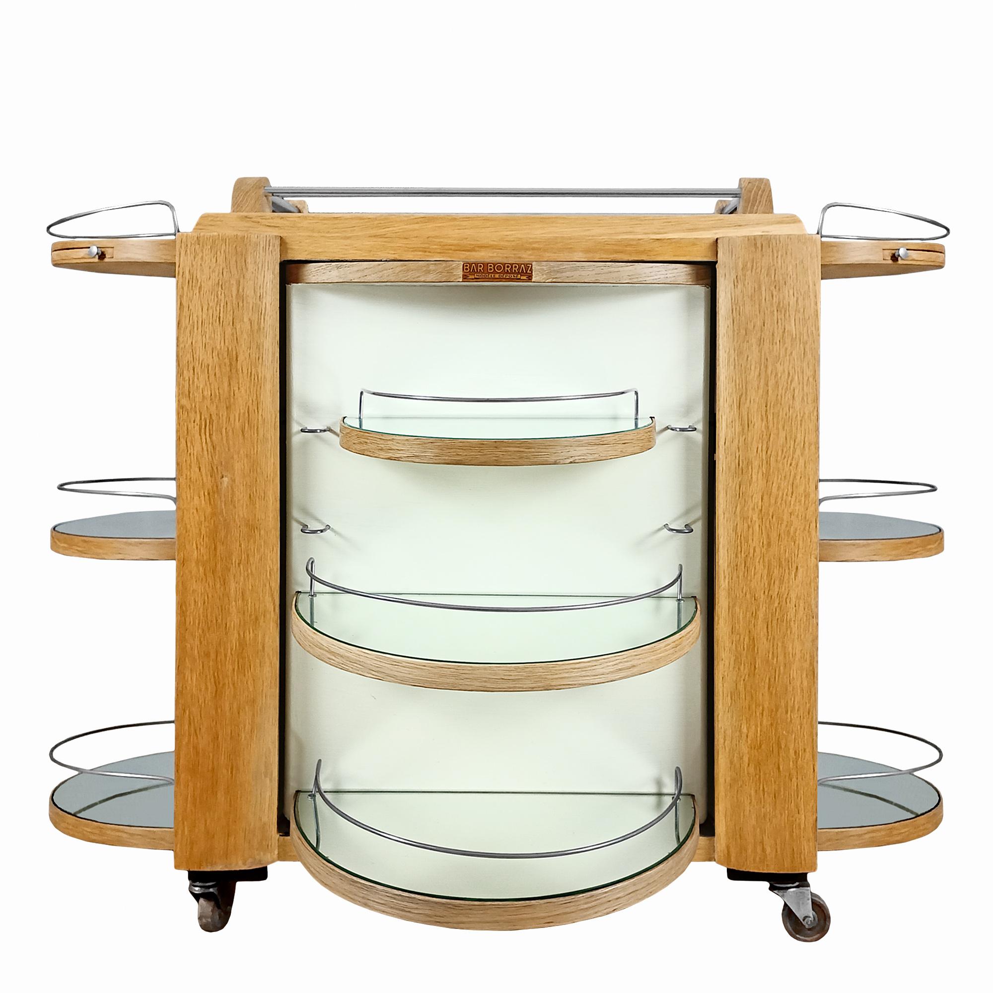 Borraz dry bar on wheels, solid oak and oak veneer, open pore finish. Pivoting door with an ivory stained space with glass and mirror shelves. Removable tray on top, two small ashtrays. Chrome plated metal hardware.

Maker’s label: Borraz registered