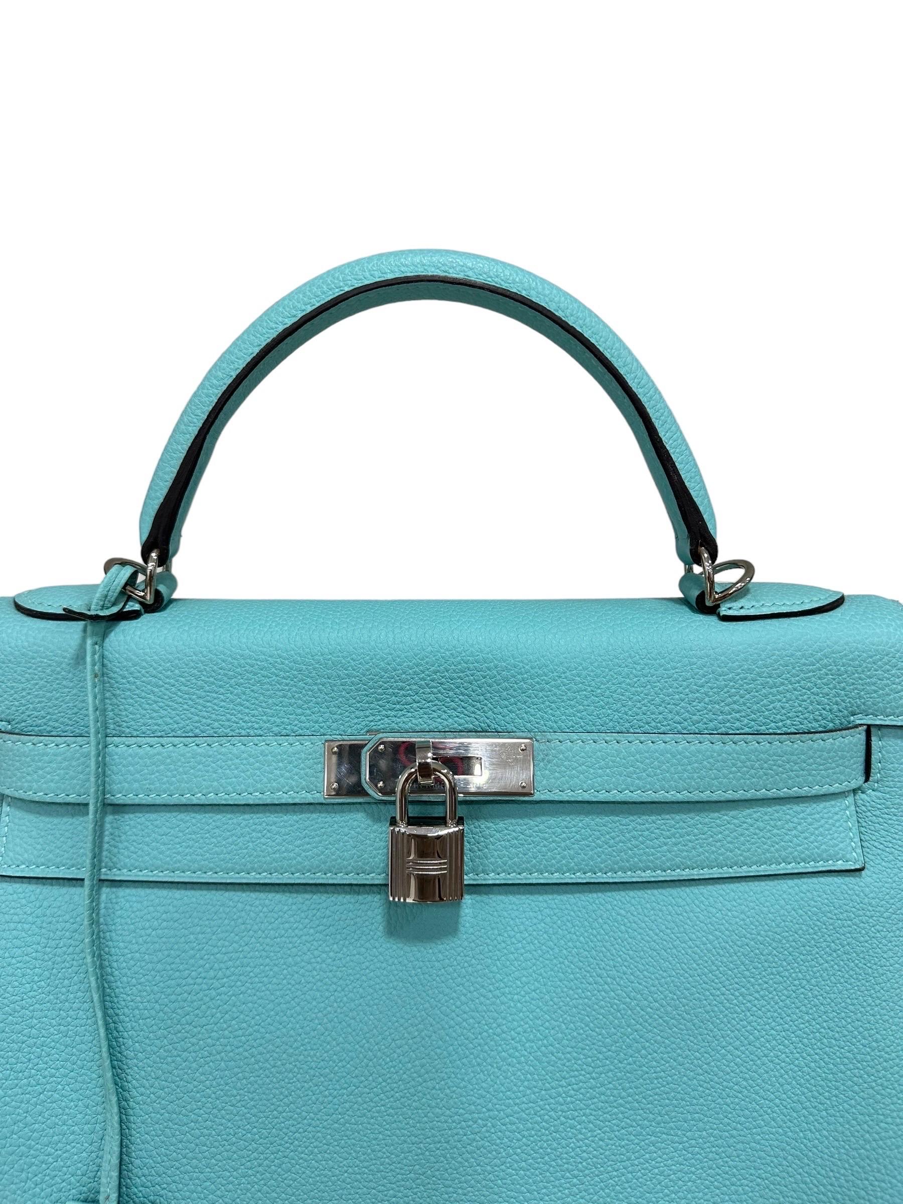 Hermès-Firmentasche, Modell Kelly, Nr. 32, hergestellt aus Pelle Clemence, morbide mit großem Granatapfel, in der Farbe Blue Atoll. Ausgestattet mit einer Klappe zum Einstecken in den Boden, mit einem Riemen und einer Schließe mit Klettverschluss.