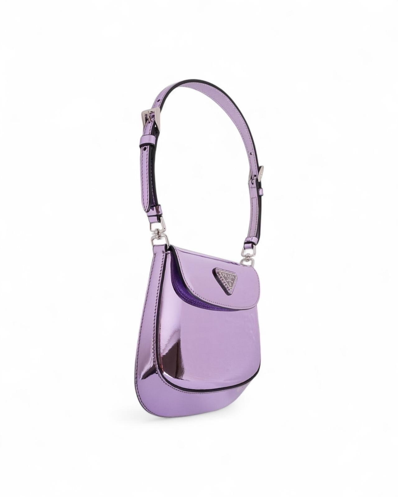 Borsa firmata Prada, modello Cleo nella misura Mini, realizzata in pelle spazzolata nella colorazione lilla con hardware argentati. Dotata di una patta frontale con chiusura a bottone calamitato, internamente rivestita in tessuto logato nero,