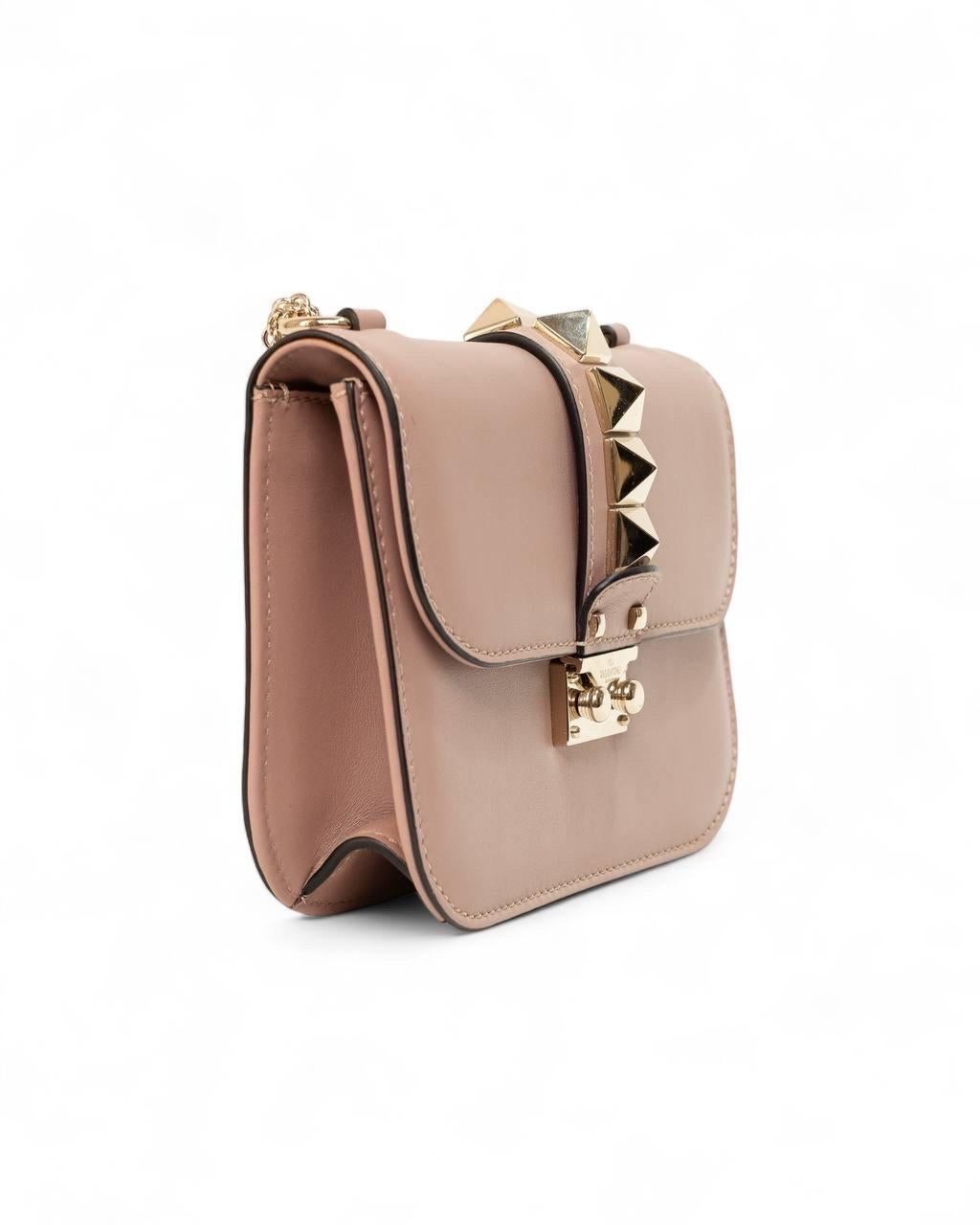 Borsa firmata Valentino, modello Glam Lock, realizzata in pelle rosa con rifiniture nere e hardware dorati. Dotata di una patta frontale con chiusura ad incastro, internamente rivestita in tela beige, capiente per l’essenziale. Munita di una