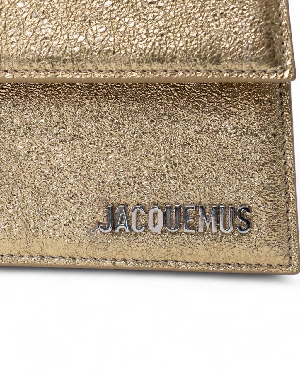Borsa firmata Jacquemus, modello Le Bambino Long, realizzata in pelle ruvida color oro metallizzato e hardware argento. Dotata di una patta frontale con chiusura interna calamitata. Internamente rivestita in tela beige, con comoda tasca, capiente