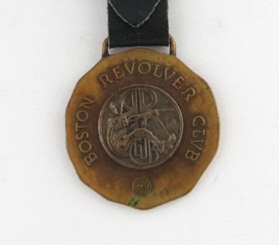 This nice watch fob and leather strap piece features a Boston Revolver Club design. The back of the bronze piece is marked Myles Standish Real Bronze NG Woods & Sons Boston. The fob is attached to a leather piece that features a gold toned buckle. 
