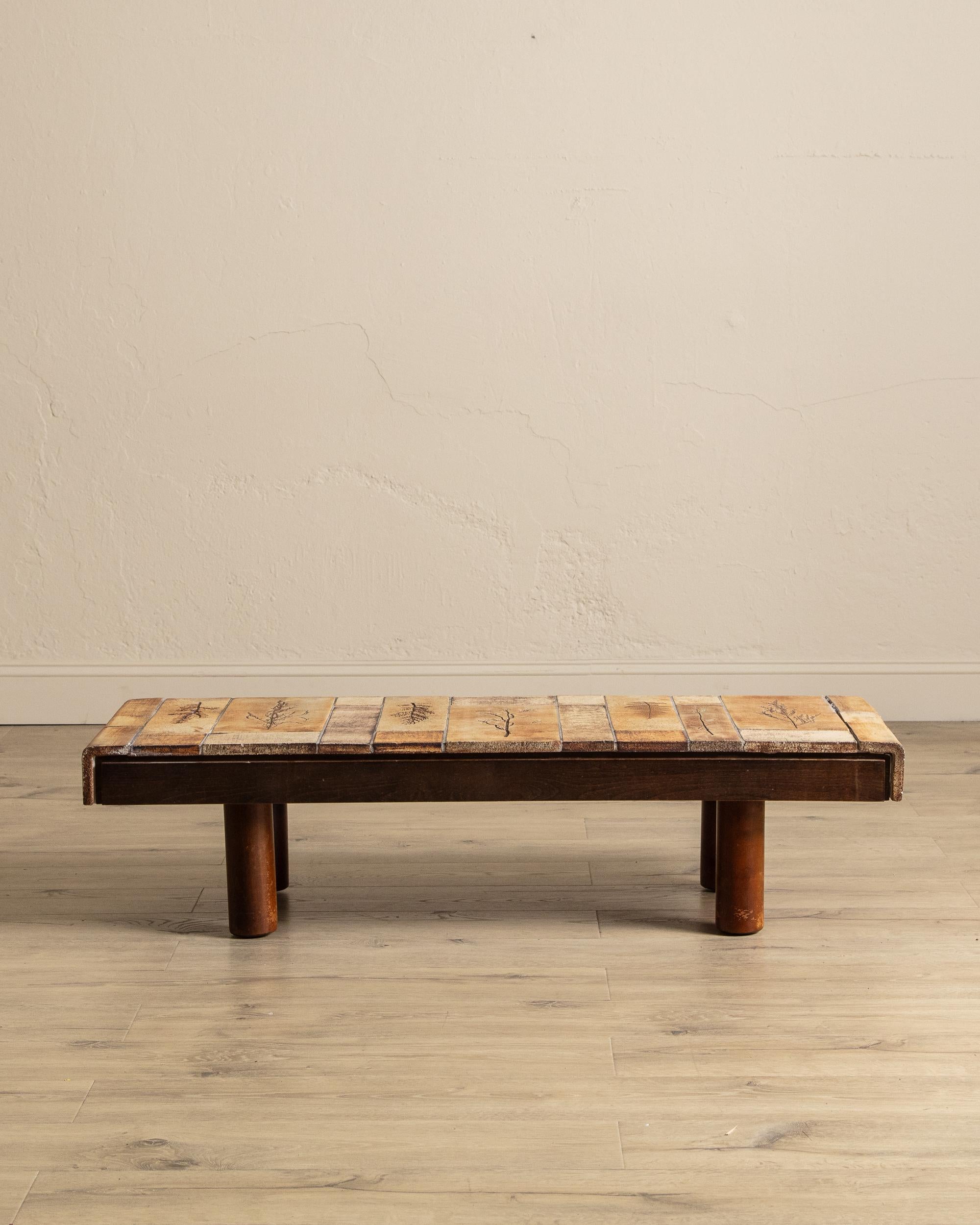 This French tile-top coffee table, signed by renowned ceramicist Roger Capron circa 1965, is a striking example of mid-century design. Its top is adorned with Capron’s signature botanical motifs, rendered in earthy, glazed terracotta tiles that