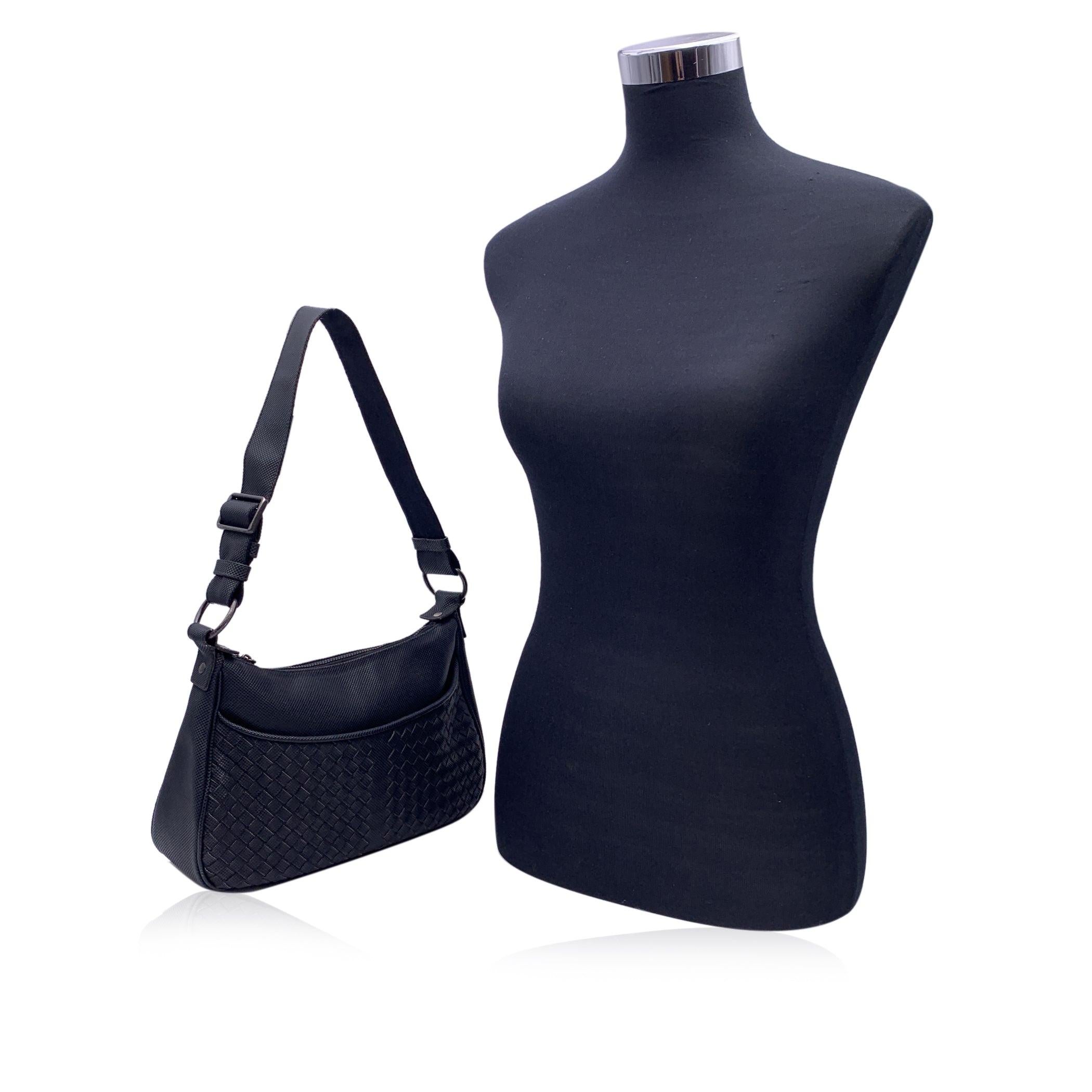Bolso de hombro Bottega Veneta de lona negra con el icónico diseño Intrecciato en la parte delantera.Herrajes de bronce de cañón. Cierre superior de cremallera.Bolsillo abierto delantero. Forro de tela negra. Correa ajustable para el hombro.