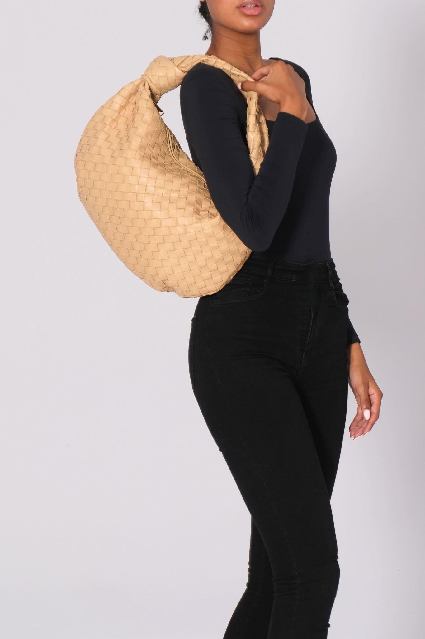 The Bottega Veneta handbag, designed by Daniel Lee, is constructed from lambskin leather in a warm beige tone. It features the signature Intrecciato weave and is complemented by gold-tone hardware. The top handle includes a signature leather knot.