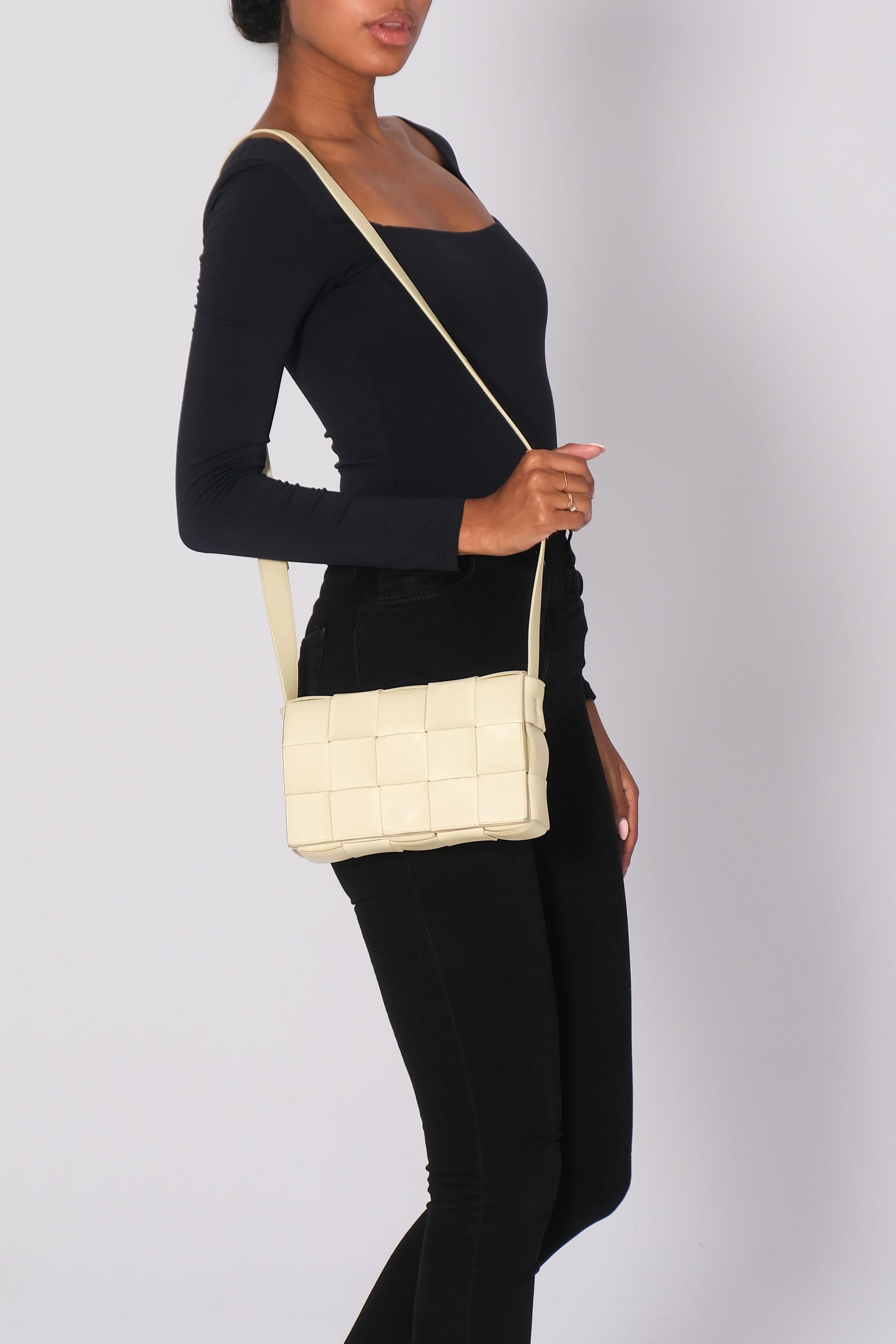 The Bottega Veneta shoulder bag is made of light yellow with the signature Intrecciato weave. It features gold-tone hardware and an adjustable flat leather shoulder strap. The exterior is characterized by its woven leather construction and a front