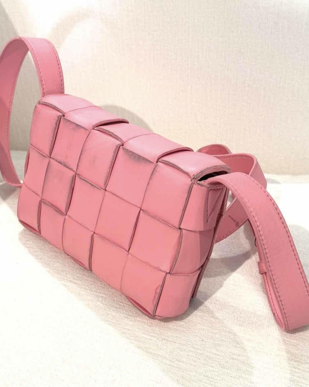 The Bottega Veneta shoulder bag is made of pink leather with the signature Intrecciato weave. The bag features gold-tone hardware and an adjustable flat leather shoulder strap. The exterior is characterized by its woven leather construction and a