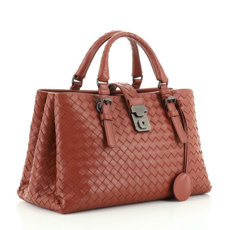This Bottega Veneta Roma Handbag Intrecciato Nappa Small, crafted in red intrecciato nappa leather, features dual woven leather handles with buckle detailing and gunmetal-tone hardware. Its push lock tab closure opens to gray suede interior divided