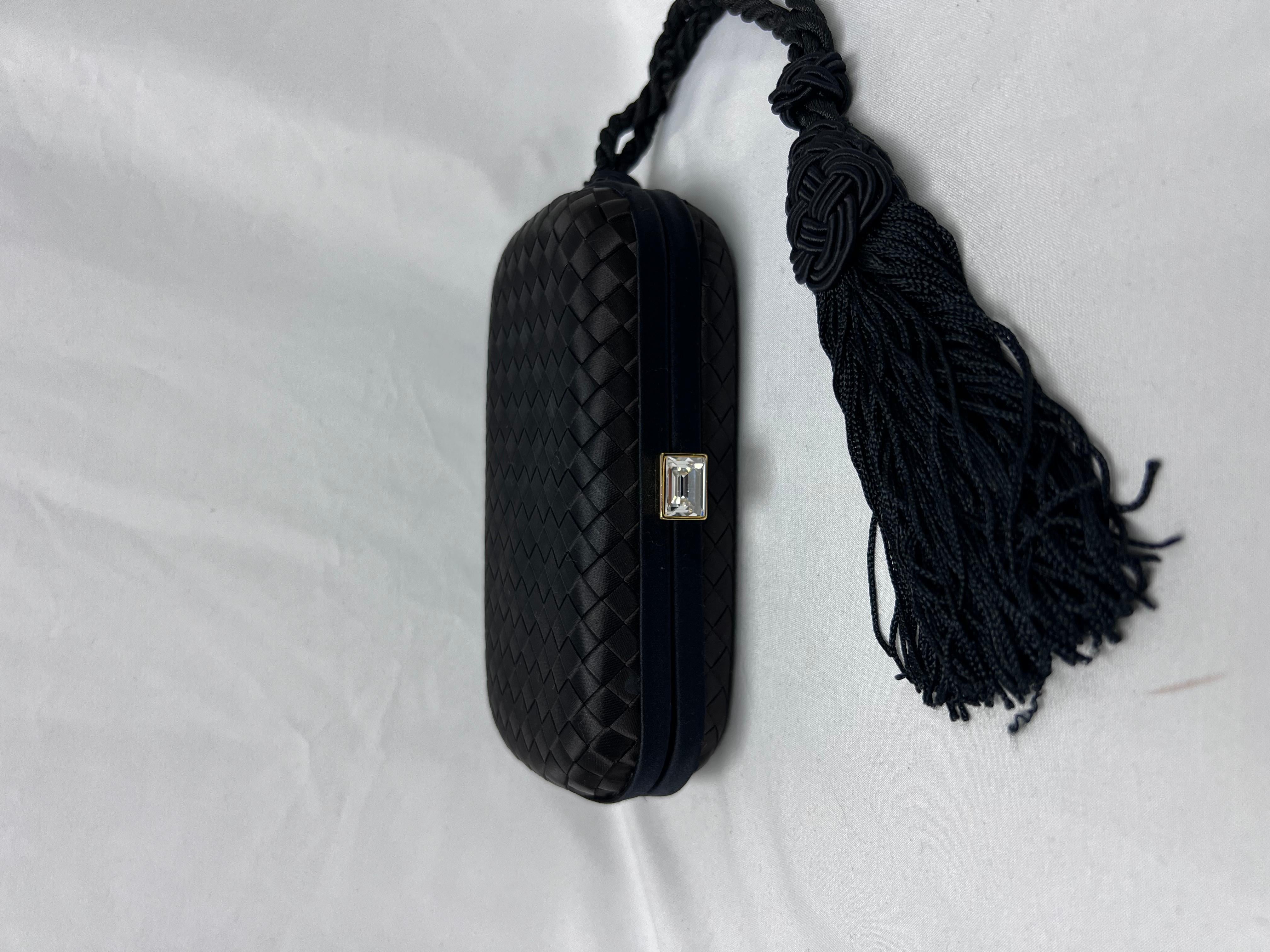The Satin Intreccio Impero Knot Clutch in Black features elegant gold hardware with a push-lock closure at the top. The stylish, structured clutch is crafted from signature intrecciato woven satin with leather trim and is finished with a distinctive