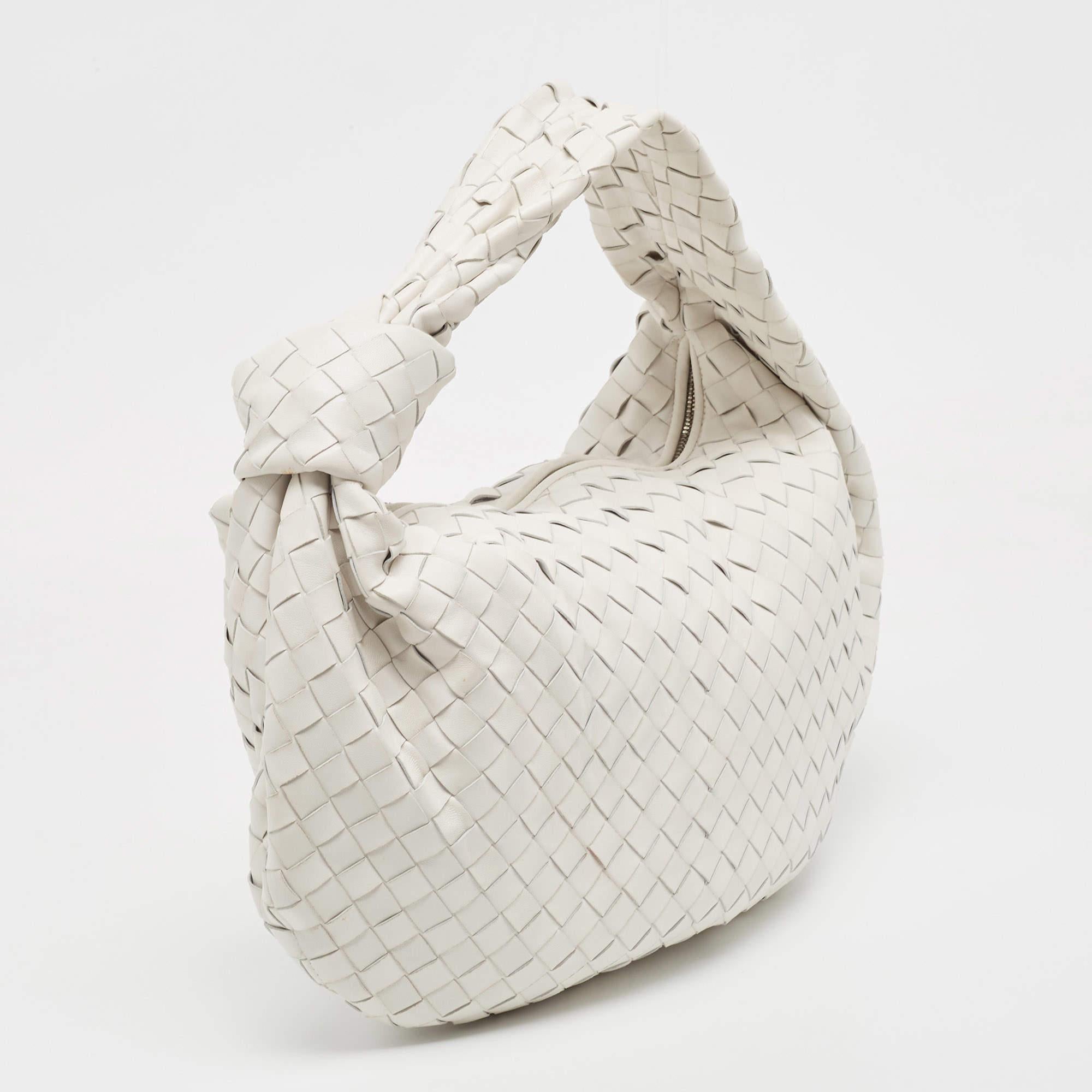 This Bottega Veneta Jodie bag is crafted from leather using their signature Intrecciato weaving technique into a seamless silhouette. This white bag, personifying elegance and subtle charm, is held by a knotted handle. The bag has an interior sized
