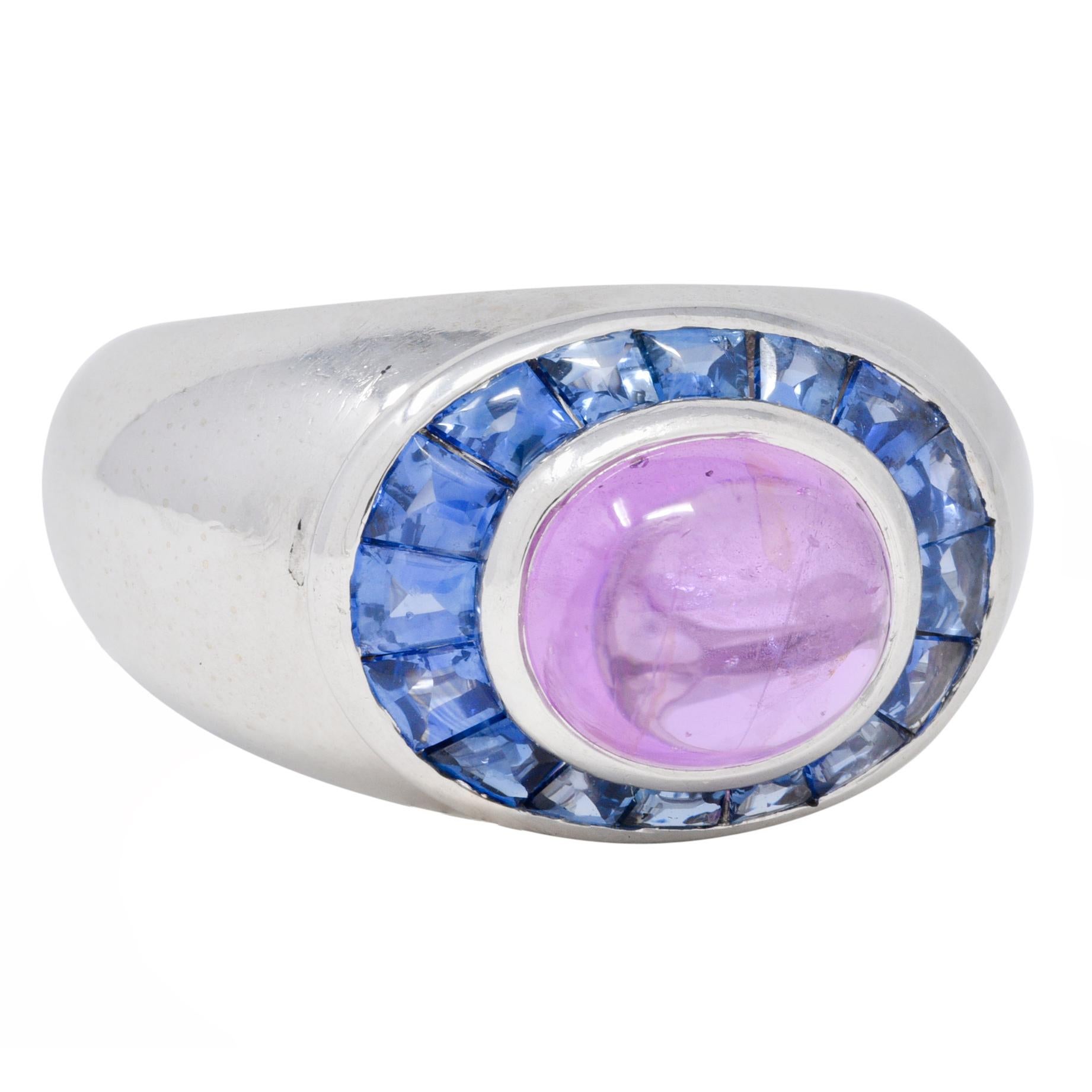 Centering an oval-shaped sapphire cabochon weighing approximately 3.82 carats total 
Transparent medium pinkish-purple in color and bezel set east to west
With a halo surround of calibré cut sapphires
Weighing approximately 1.26 carats total