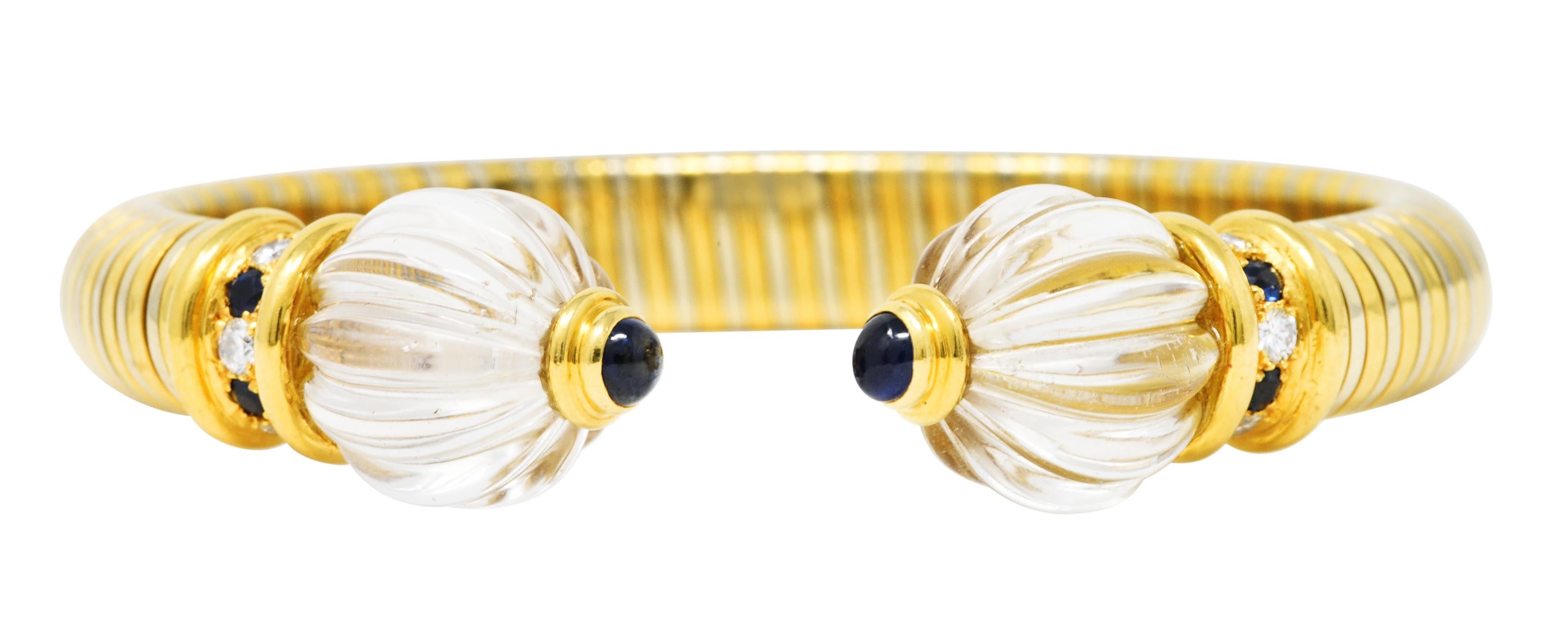 Cuff bracelet is designed as a white and yellow gold flexible tubogas with 14.0 mm round rock crystal quartz terminals. Carved with deeply grooved segments and featuring bezel set 3.5 mm round sapphire cabochons. Accented by round cut sapphires