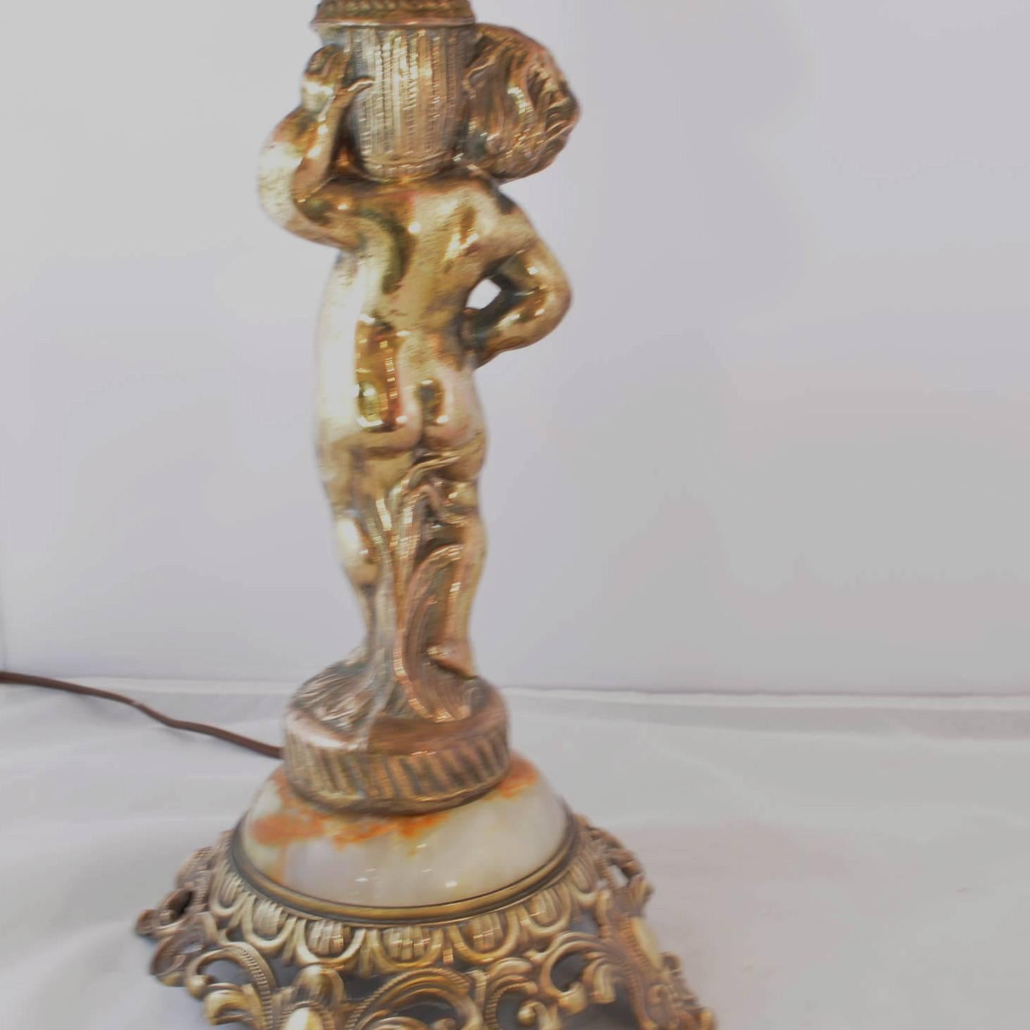 The putti holds an urn on his shoulder and sits atop a base of domed marble and cut metal base. When lit he has a warm golden glow. The charming shade has alternating panels of lace and gathered fabric with rosette centres. 

Dimensions: H 23