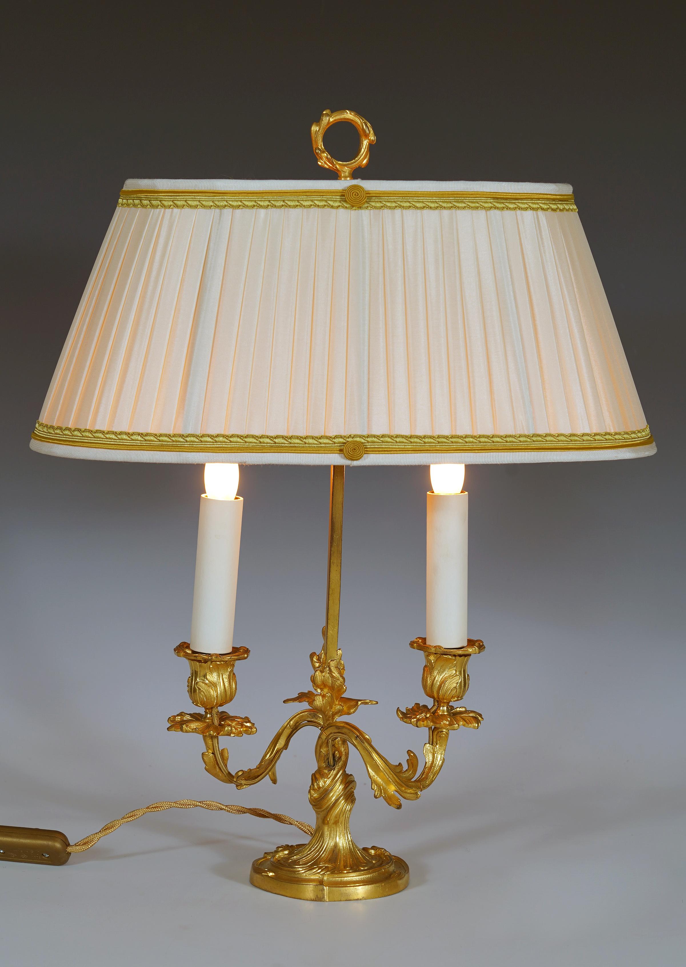 Encantadora lámpara bouillotte de inspiración Luis XV, de bronce cincelado y dorado, con dos brazos de luz decorados con follaje, rematada con una elegante pantalla ovalada de seda plisada beige decorada con trenza dorada.

Las lámparas Bouillotte