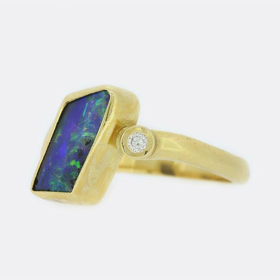 This is an 18ct yellow gold boulder opal and diamond abstract ring. The central opal is rub-over set, naturally shaped and has a wondrous play of colour. This focal stone is complemented by a single round cut natural diamond which sits harmoniously