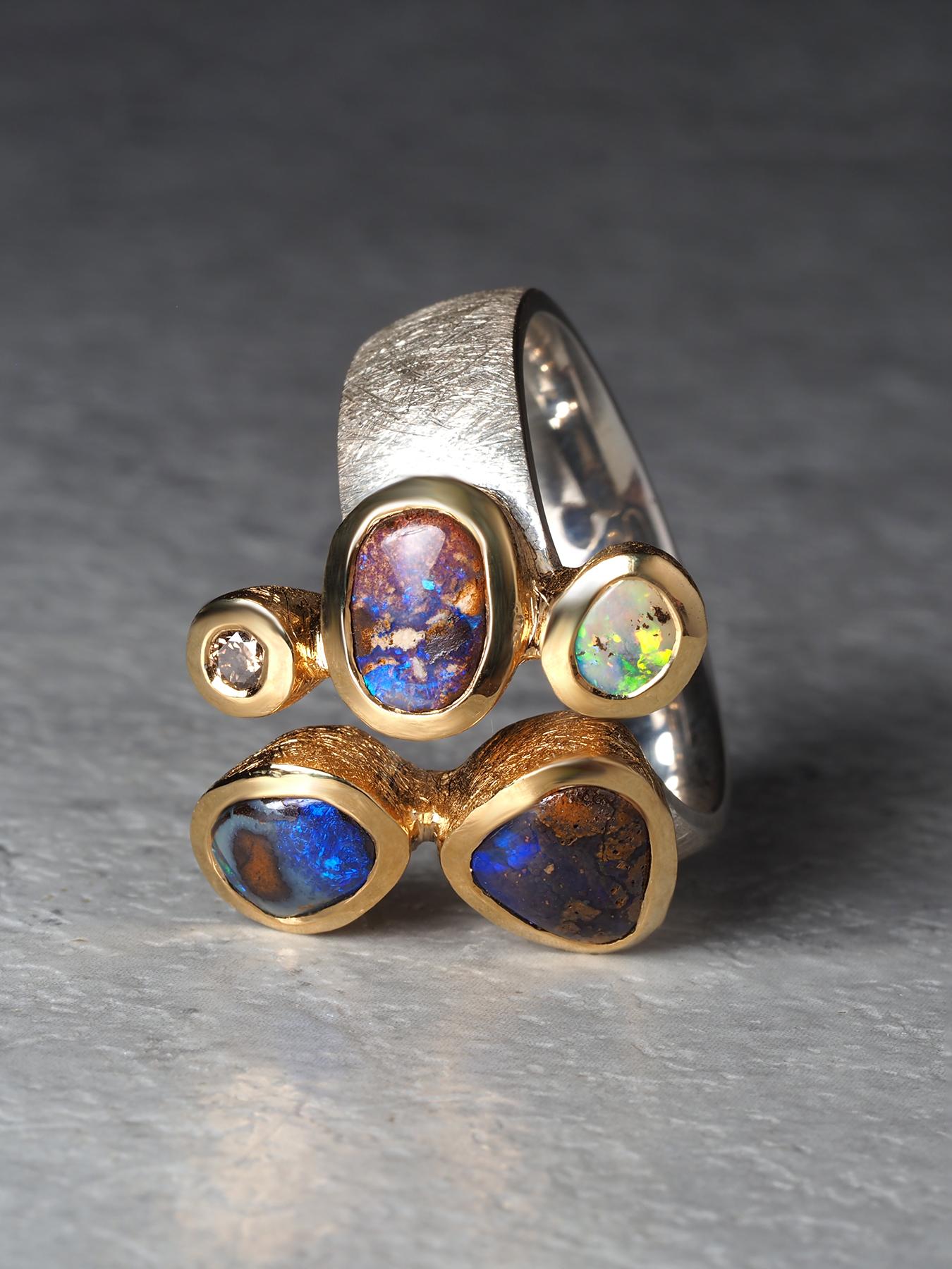 Ein skulpturaler Ring mit vier natürlichen australischen Boulder-Opalen und einem einzelnen runden Diamanten. Die Opale zeigen ein auffälliges Zusammenspiel von tiefen Blau- und Grüntönen mit violetten und roten Reflexen, wobei jeder Stein sein