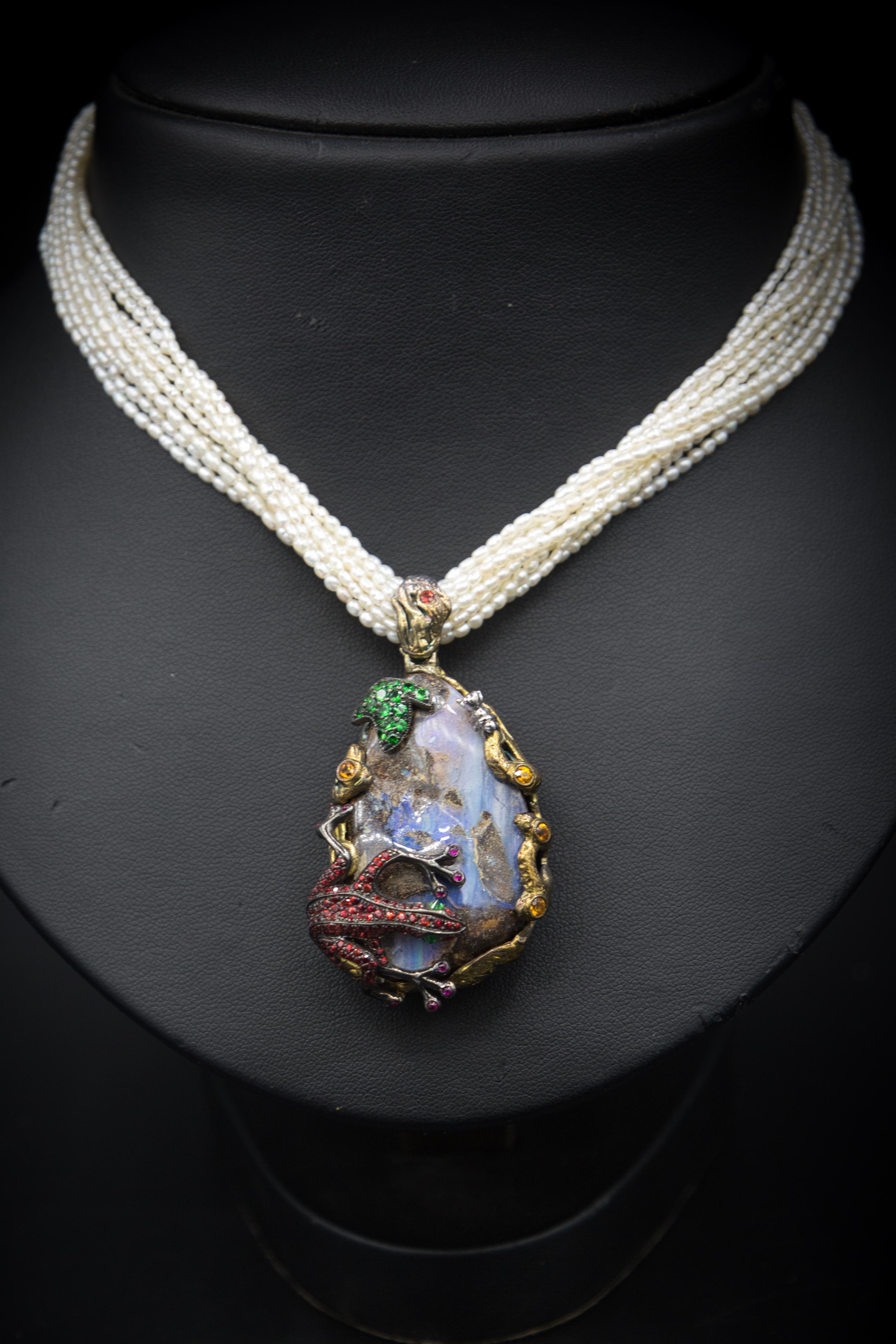 Il ciondolo opale è un gioiello artistico che unisce eleganza e natura. Il ciondolo presenta un imponente opale boulder ornato da una rana d'argento con zaffiri rossi brillanti. Una foglia di tsavoriti e zaffiri parzialmente arancioni completa il