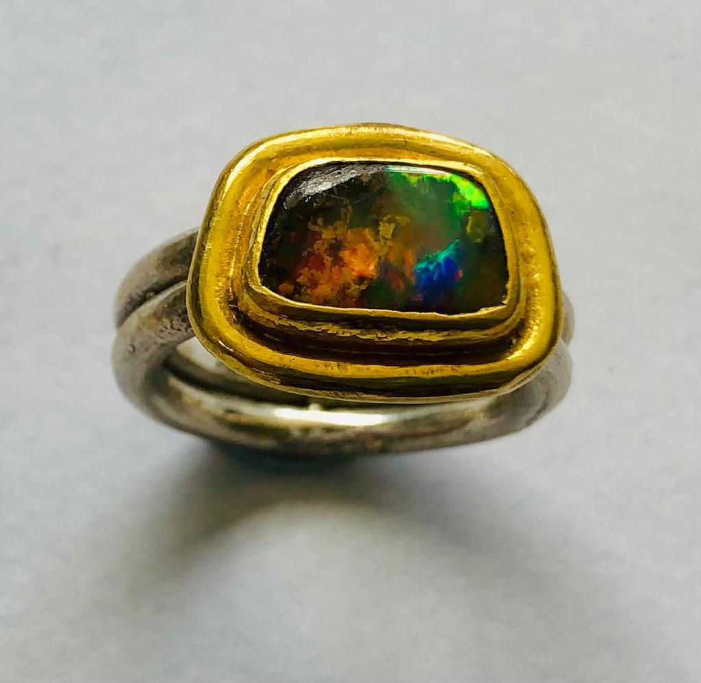 This beautiful handmade boulder opal ring has an array of different colours and lights including flashes of red, blue and green. Set and framed in rich, textured 22 karat gold, this stunning opal sits on a double band of sterling silver and the