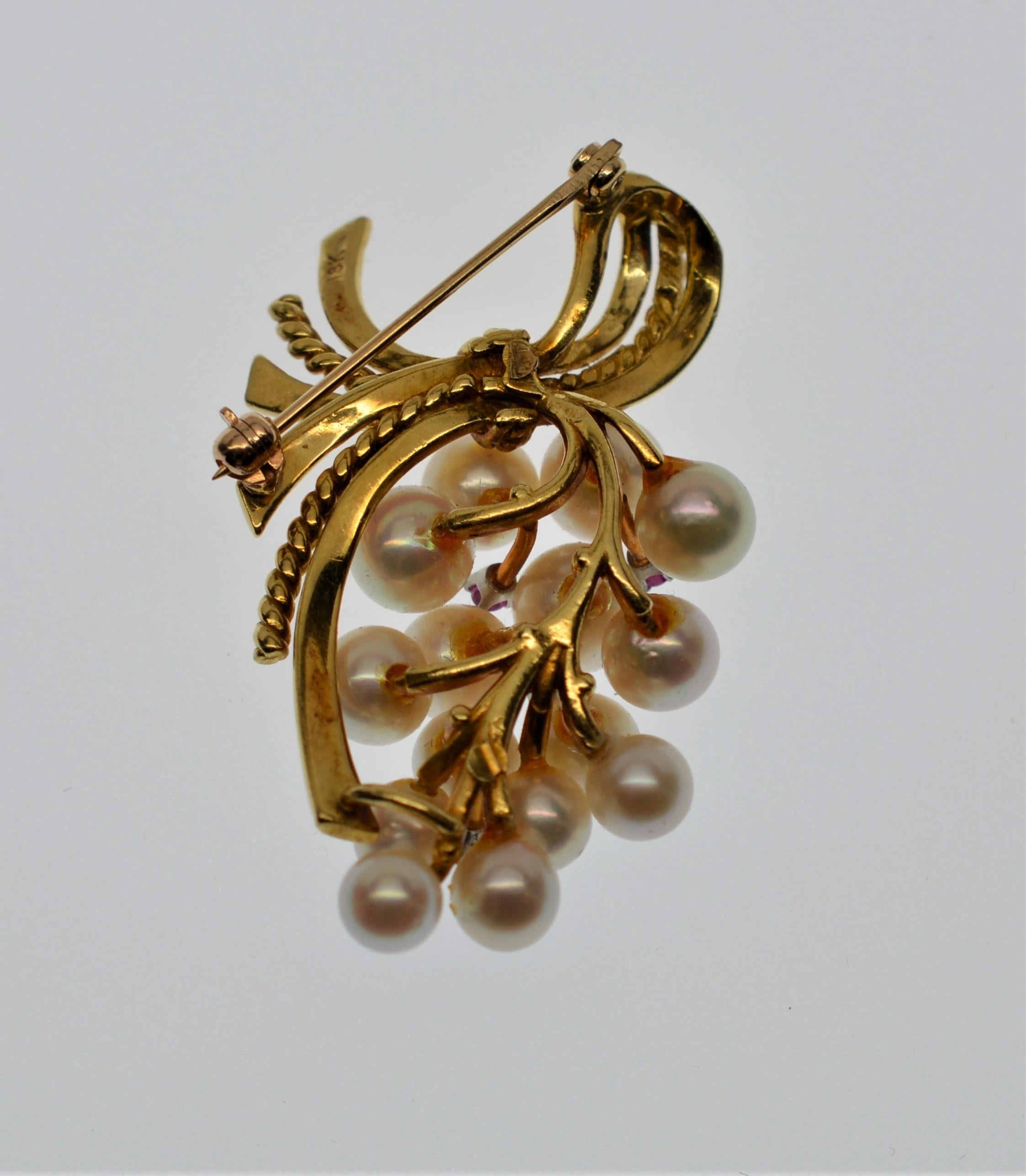 Enjoy this lovely late 1950's eighteen karat (18K) yellow gold brooch with a cascading cluster of lustrous Akoya pearls highlighted with ruby accents artfully gathered by satin eighteen karat gold ribbon.  Attractively arranged AAA pearls range in