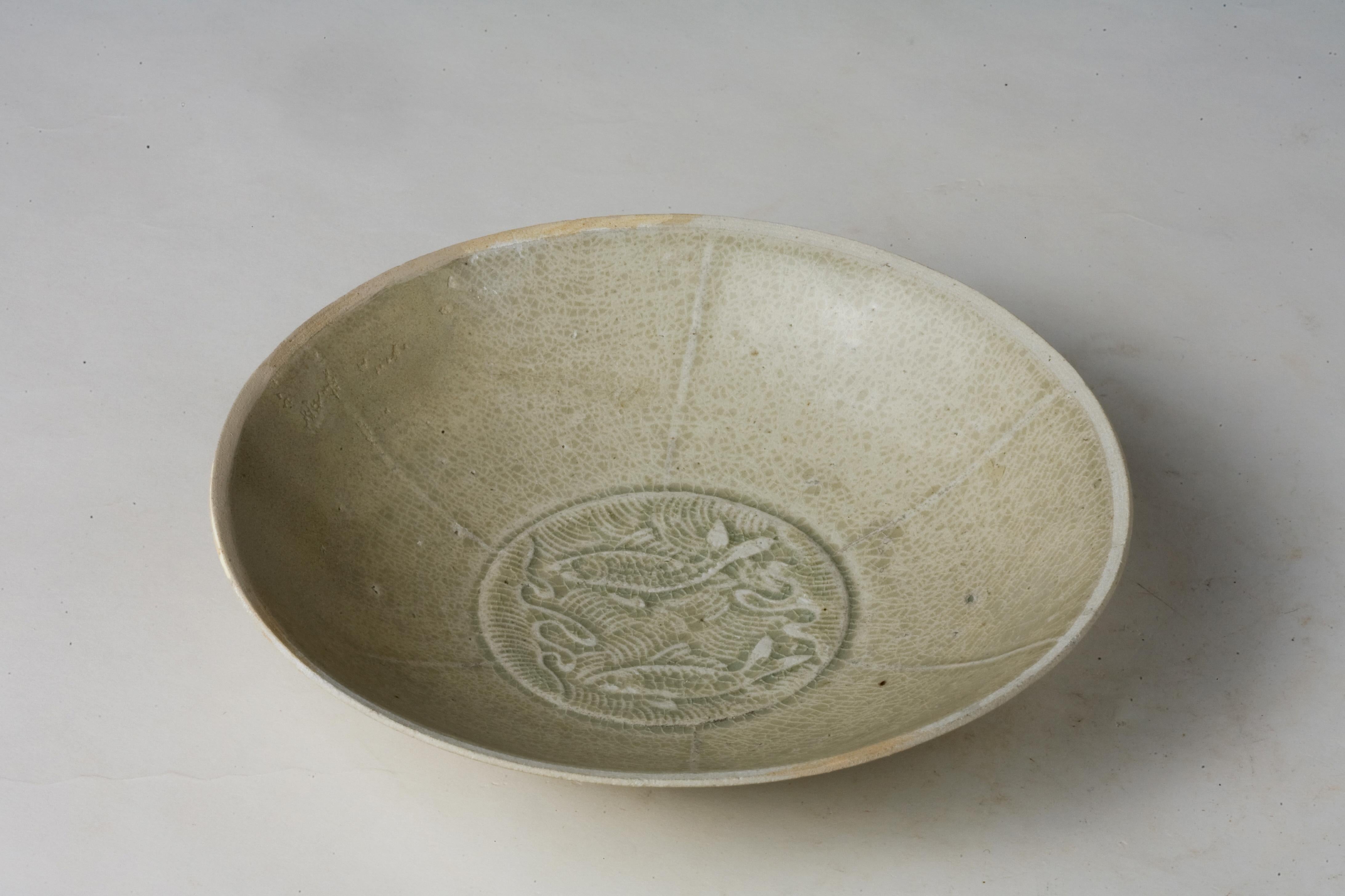 El cuenco tiene un borde ancho y delgado y un pie bajo, característico de las formas de la dinastía Song. Un diseño de dos peces finamente tallados y llenos de vida decora el interior, un motivo auspicioso clásico de la cerámica Qingbai. El vidriado