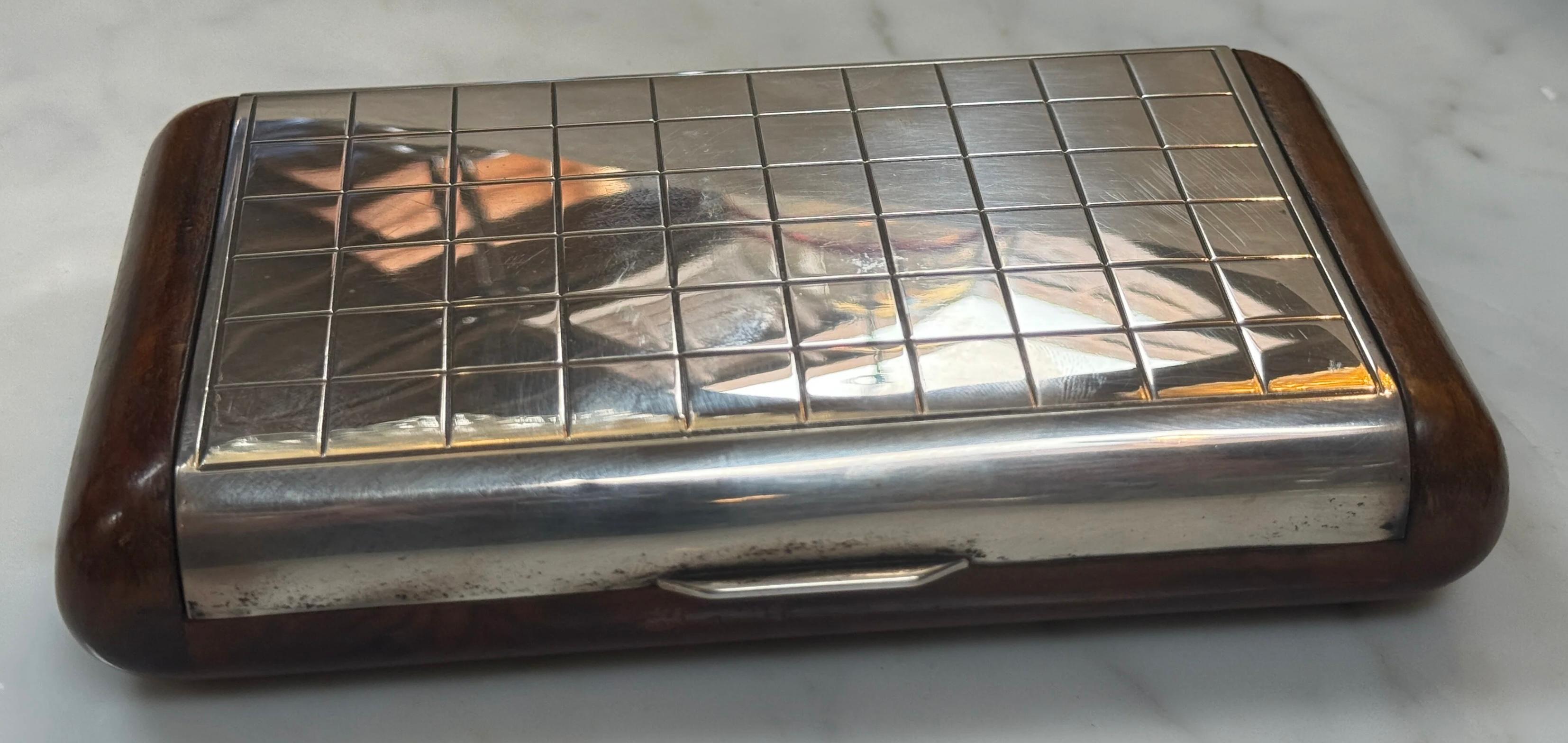 Es handelt sich um eine kleine rechteckige Kiste mit einem Metalldeckel mit Gittermuster und Holzseiten. Sie hat einen Riegel an der Vorderseite und scheint für kleine Gegenstände wie Zigaretten oder Karten verwendet zu werden.
