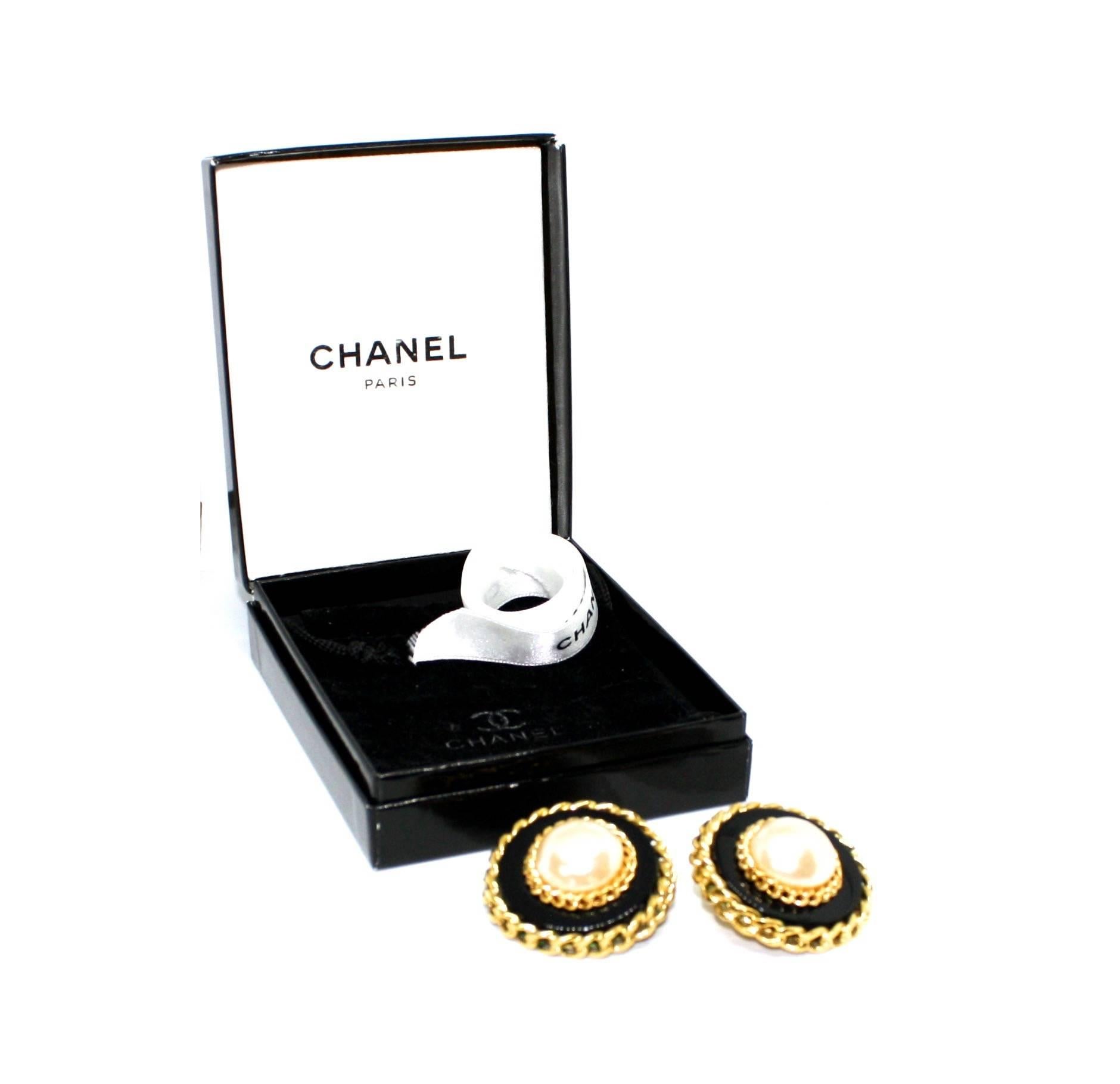 Iconic clip earrings by Chanel featuring a large central pearl surrounded by black acrylic and gold plated chains. Dated to the late 1980's/early 1990's, each earring is stamped with the Chanel Cartouche. The clip earrings are also provided with the