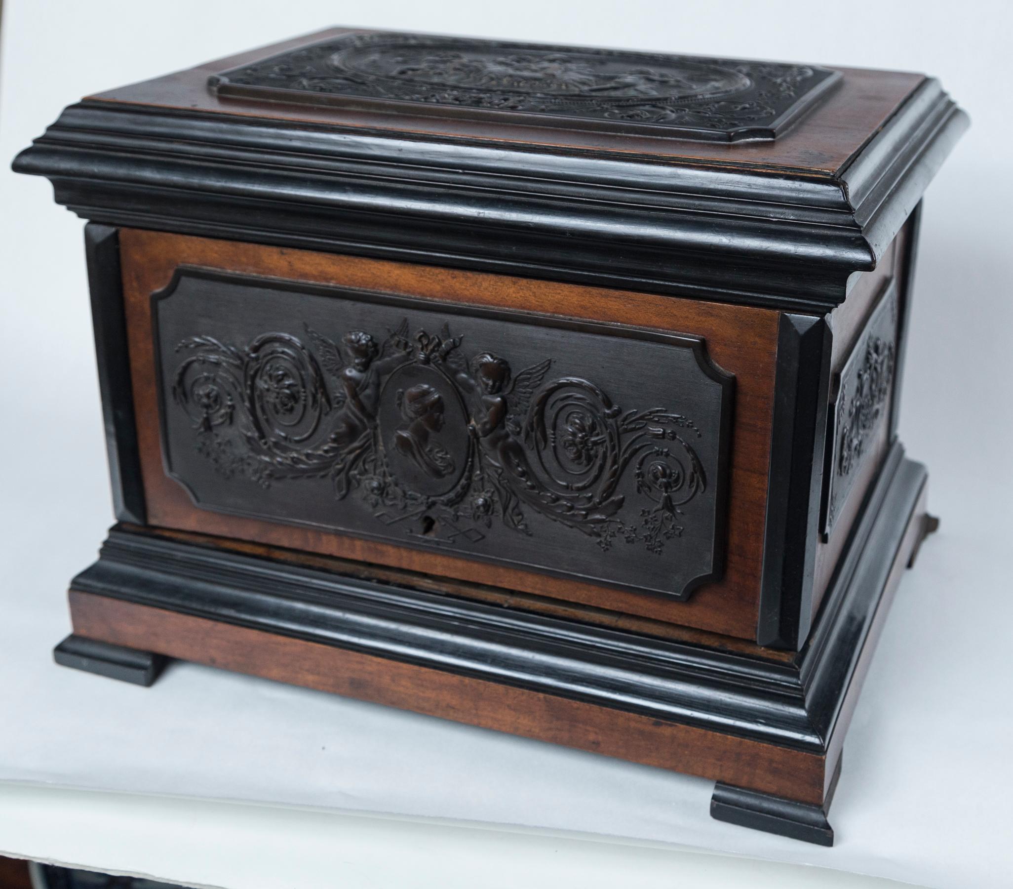The box of a well grained polished mahogany or other hard wood with ebonized moldings and feet, applied dark bronze plaques of cavorting putti with a goat (top) a classical medallion of a female profile with putti on either side (front) and panels