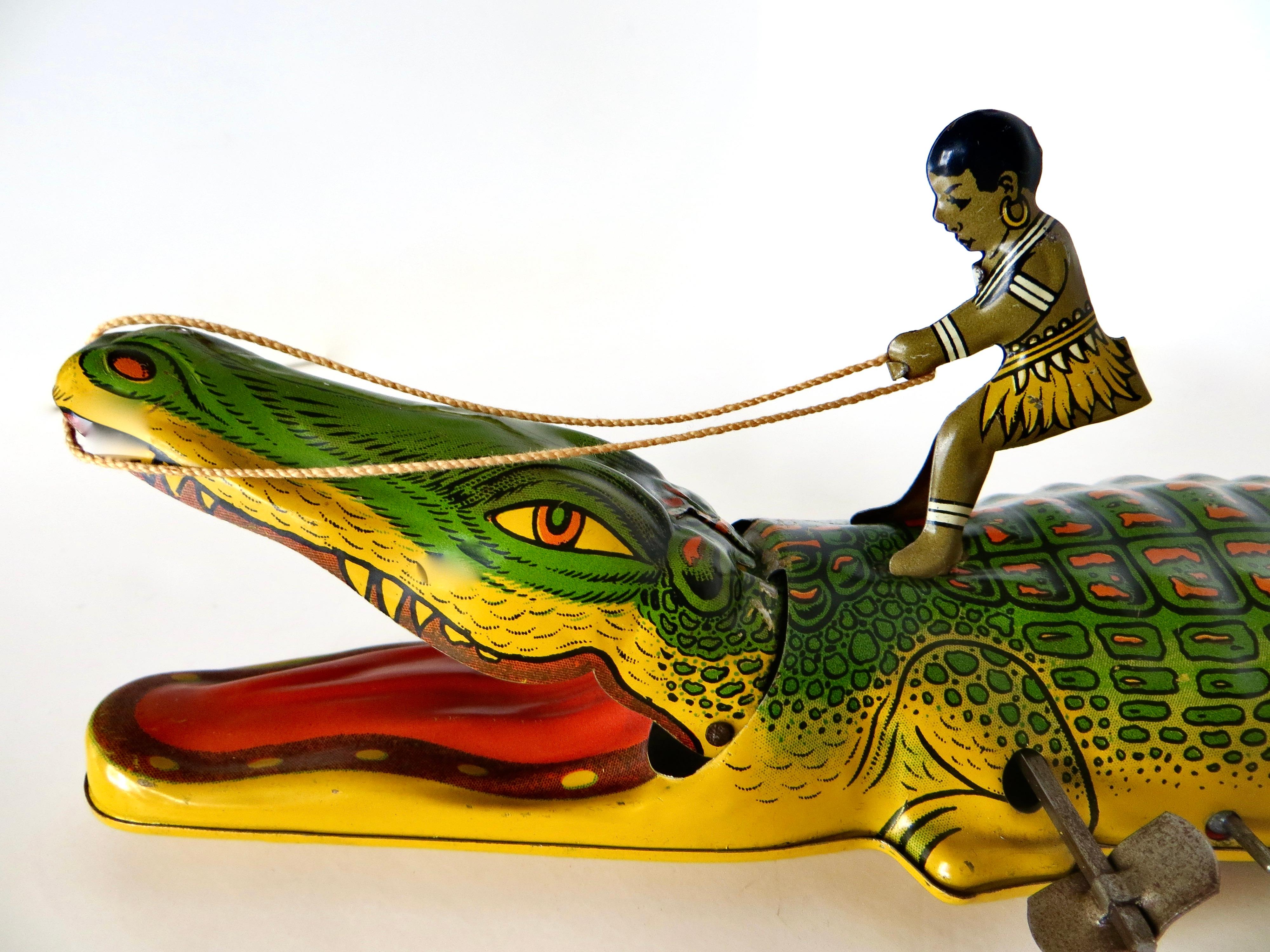 Dieses Vintage-Blechspielzeug, das um 1935 von der J. Chein Company in Harrison, New Jersey, hergestellt wurde, zeigt im aufgezogenen Zustand einen kleinen Jungen in Insulanerkleidung, der versucht, auf einem Alligator zu reiten, der über den Boden