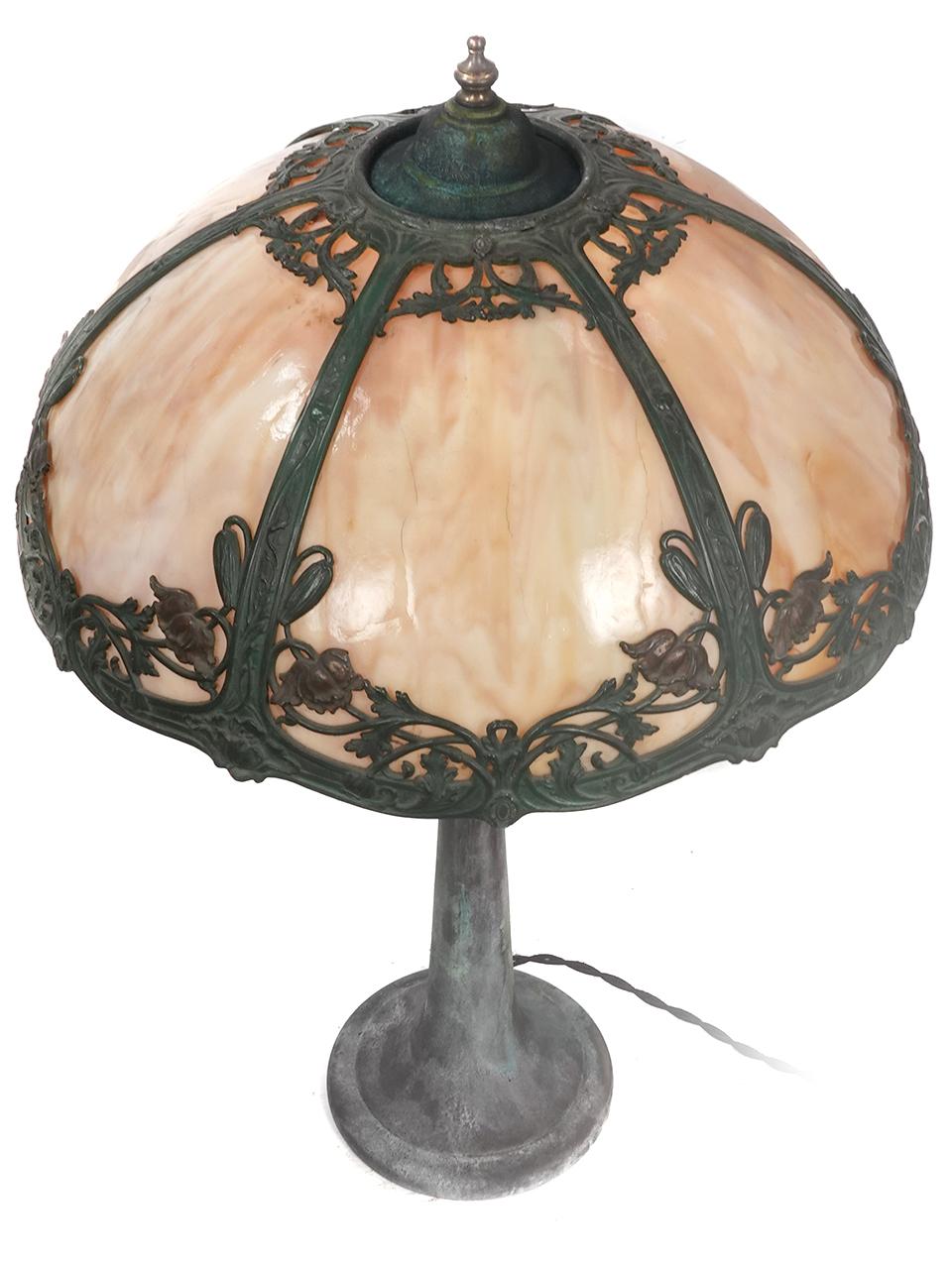 This Art Nouveau table lamp is in the manner of Bradley & Hubbard and offers a slag glass paneled shade with filigree floral details. The glass gives off a nice warm glow.