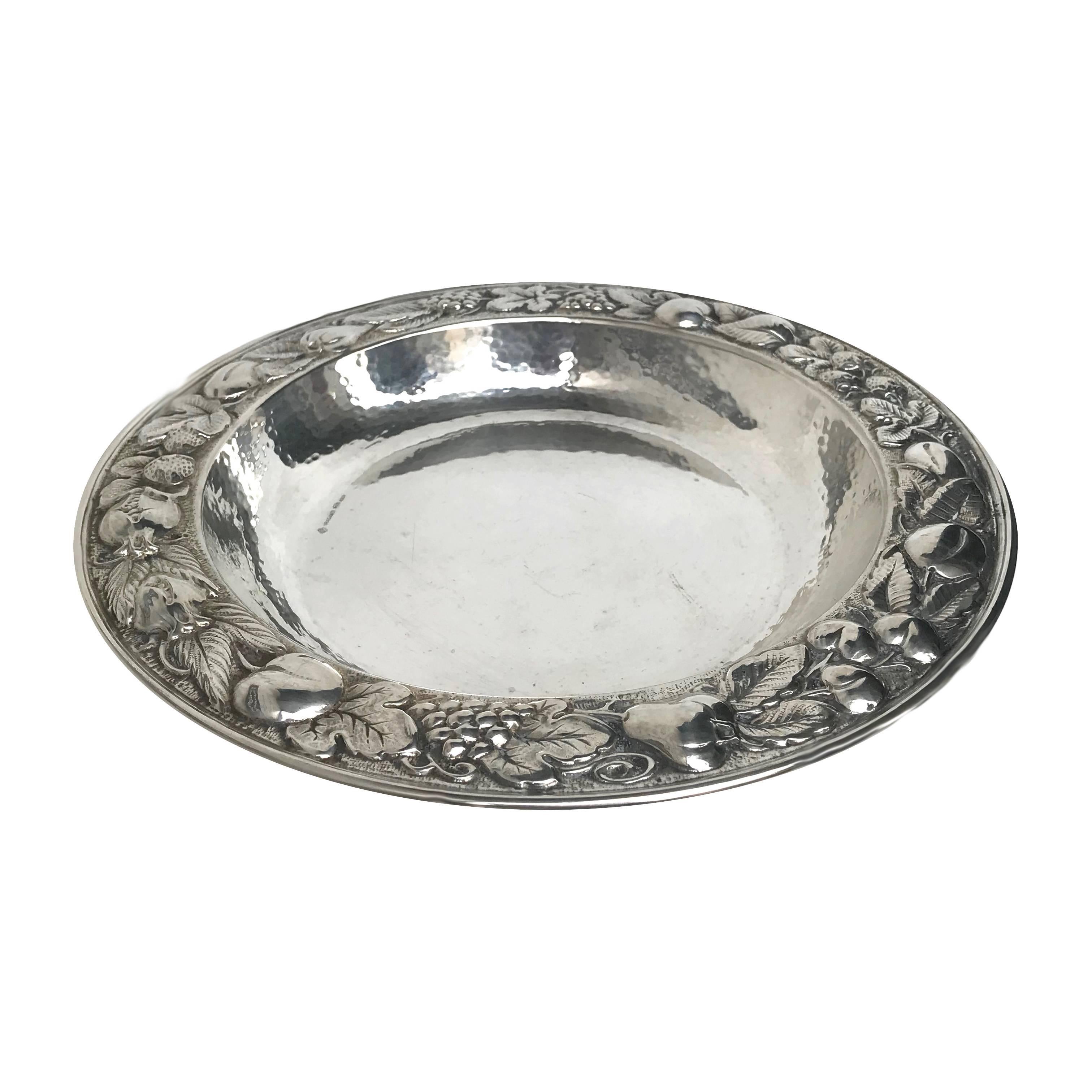 This elegant centrepiece is a deep dish entirely made in silver whose internal surface is textured with the classic hammered surface, while the lip is intricately adorned with a magnificent chiselled decoration that depicts a garland of leaves,