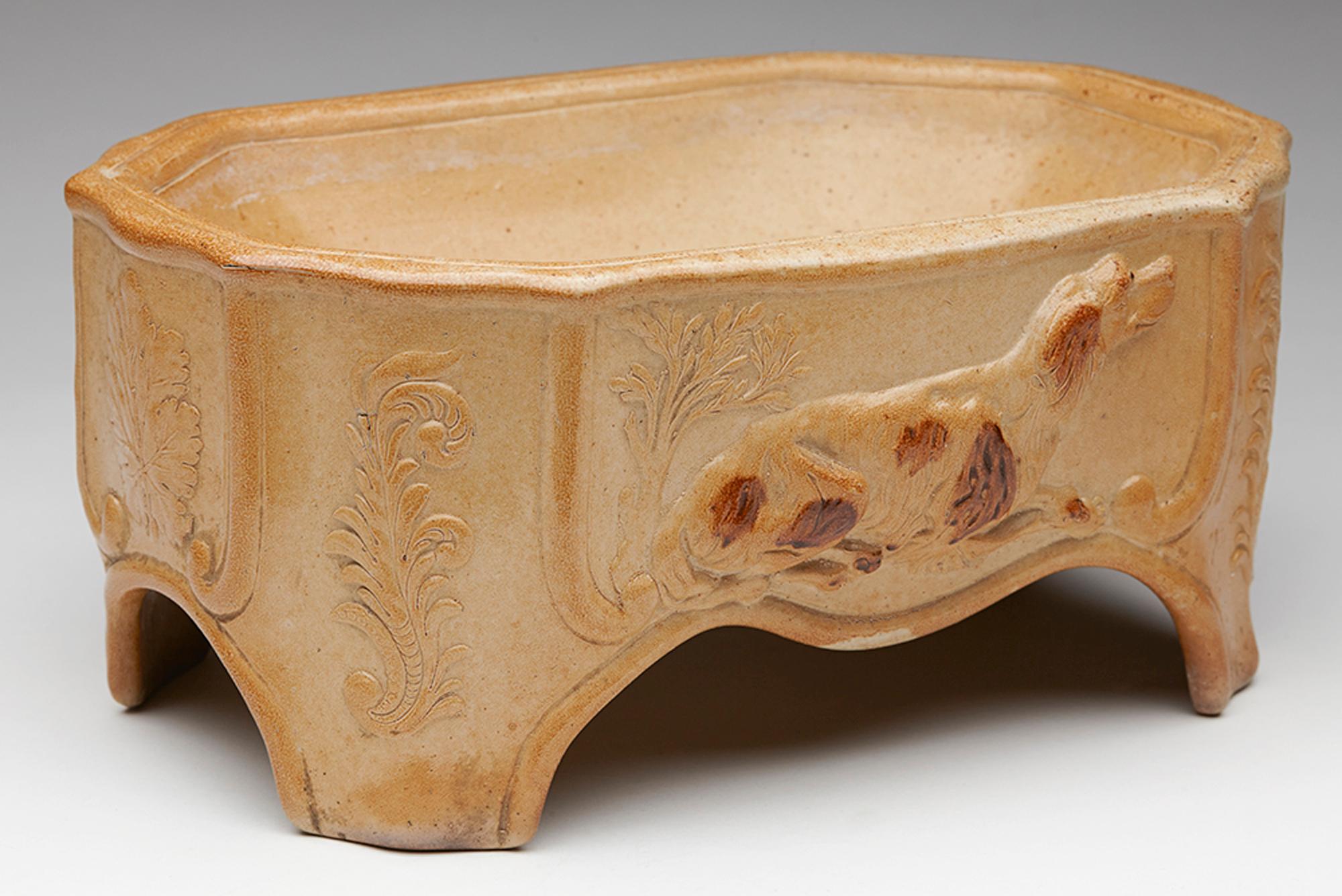 A rare antique English, Brampton salt glazed dog bowl molded with setter like dogs and leaves and dating from the mid 19th century or possibly slightly earlier. The eight sided stoneware bowl is of rectangular shape and stands raised on four corner