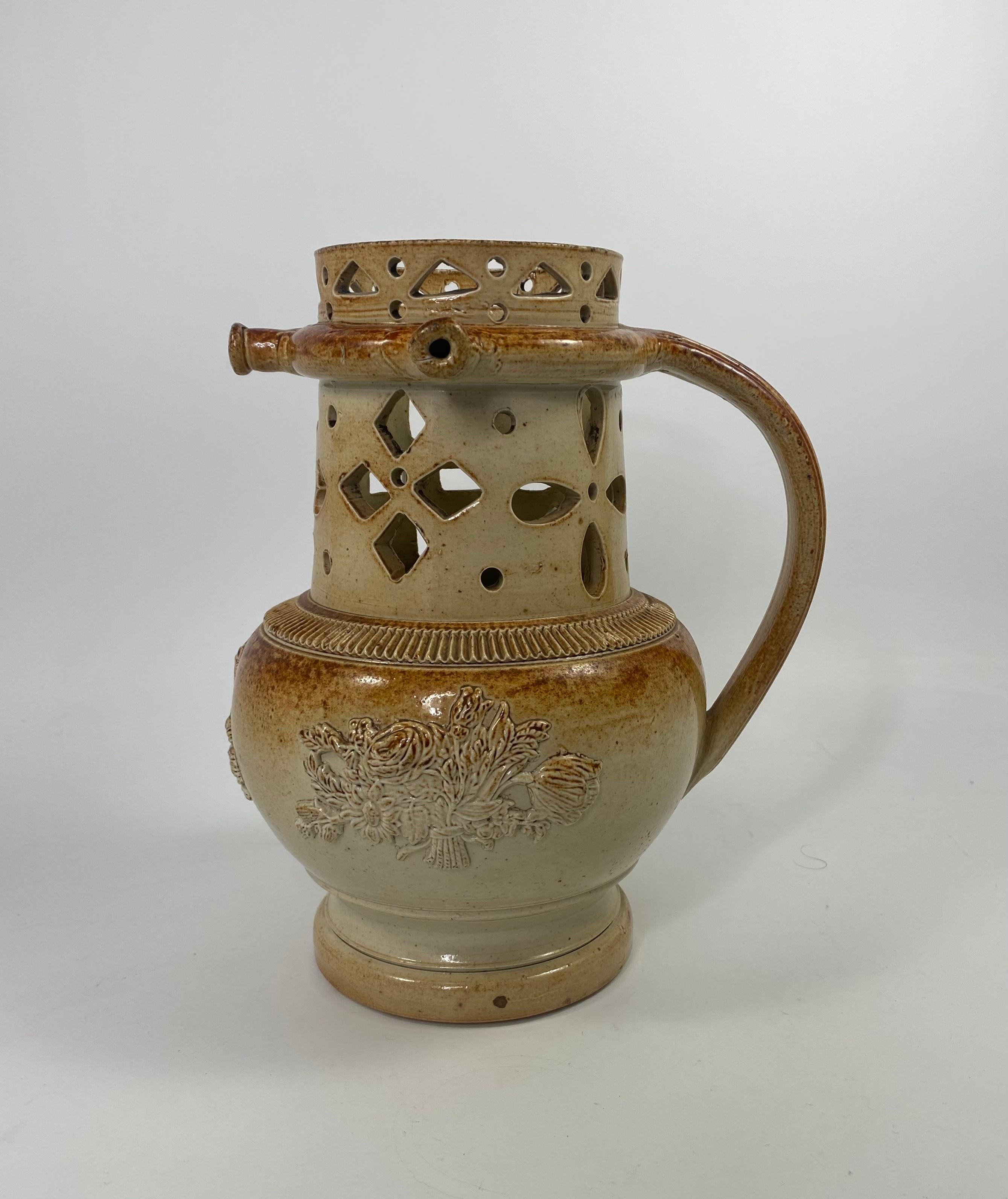 A fine and large Brampton saltglazed stoneware Puzzle Jug, c. 1840. The jug sprigged to the body, with reliefs of musical instruments, a spray of flowers and a windmill. Having three spouts, which are connected to the hollow handle. The neck