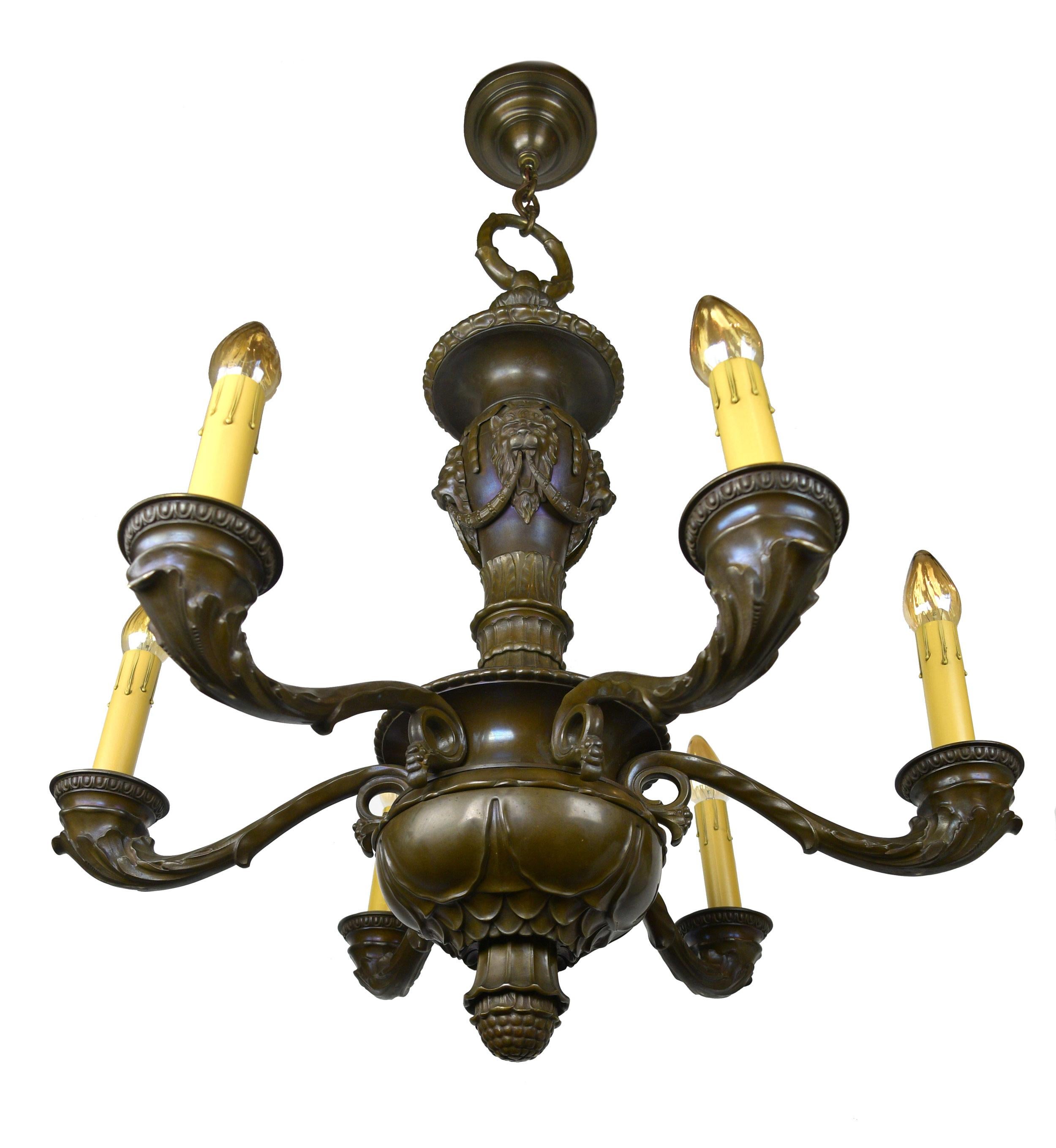 This cast bronze six candle chandelier has a grounding presence. Acanthus leaf arms curl at the body and flow out to hold the detailed basins of the candle holders. The lion quadrifrons are connected by Laurel wreaths with berries, both of which are