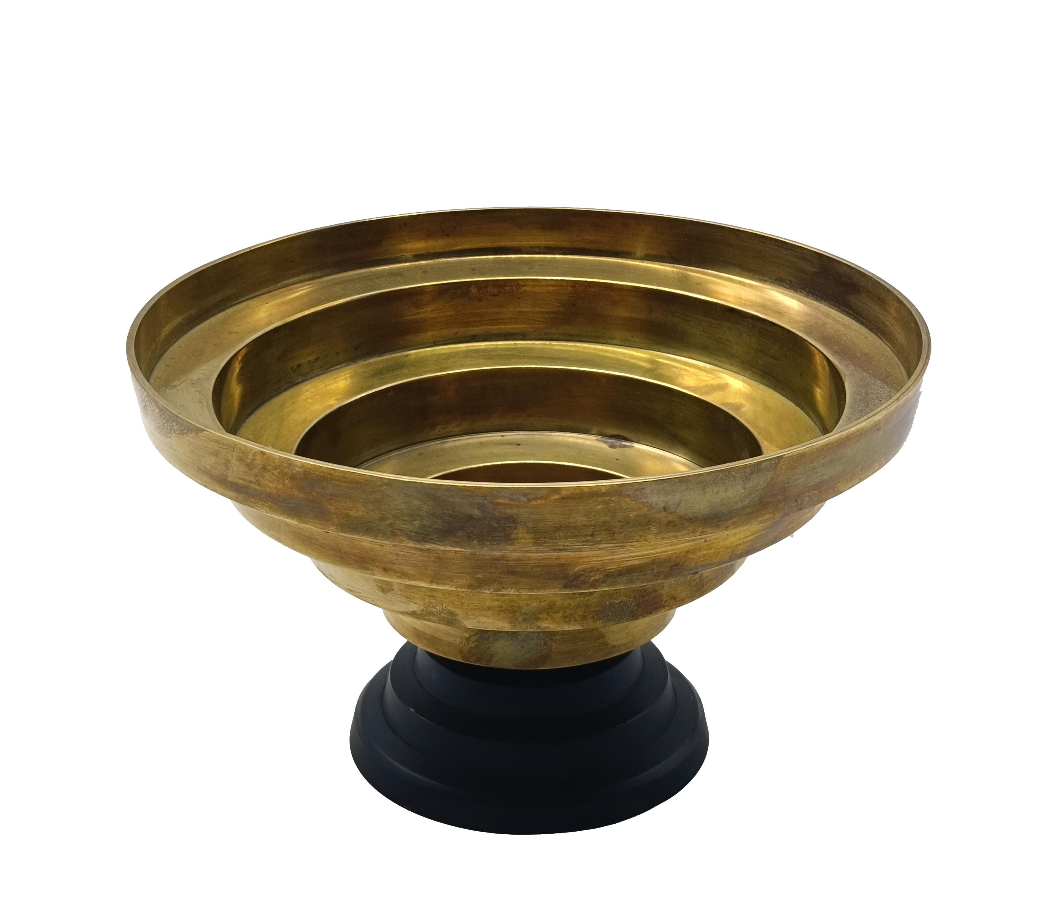Grazioso centrotavola forma a coppa Art Deco' in ottone e base laccata nera. Questa raffinata coppa si distingue per il design scultoreo e la finitura in ottone spazzolato, che diffonde riflessi caldi e sofisticati nell'ambiente. Le linee essenziali