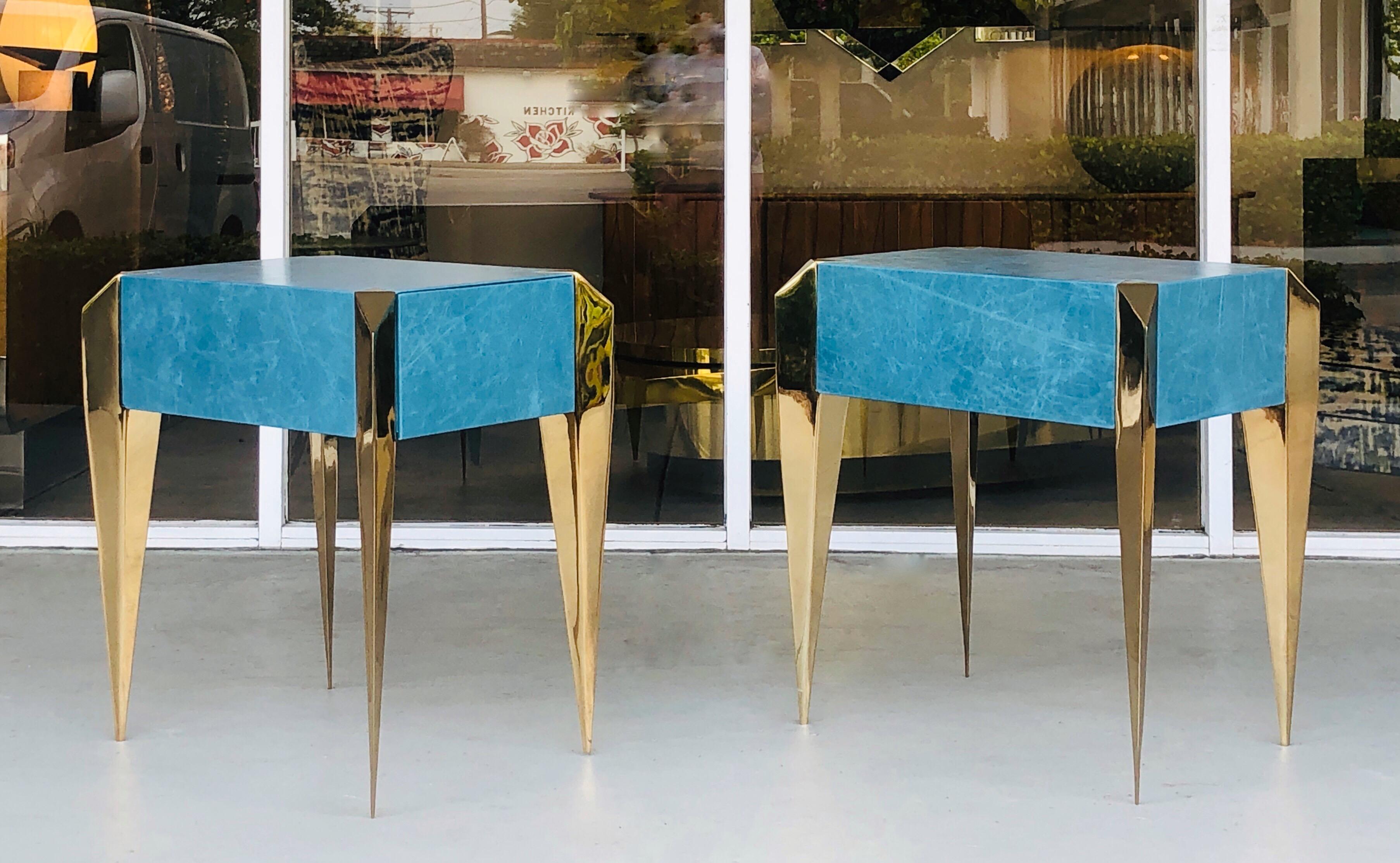 A pair of modern sculptural side tables. The body is clad in cerulean blue leather, the prism like legs are gold plated metal. One-drawer each.
