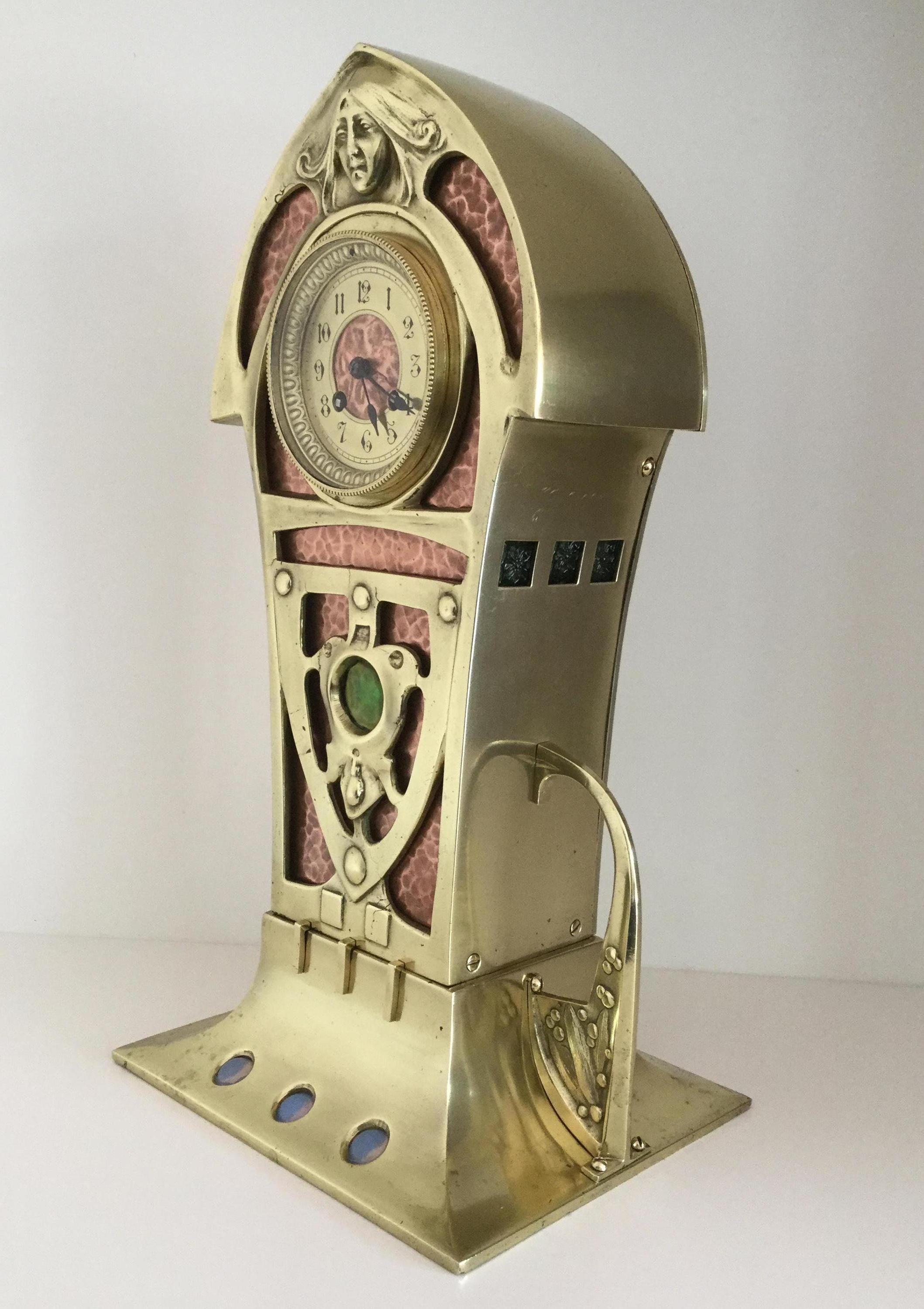 A stunning large Art Nouveau brass and hammered copper mantel or table clock, the pointed arch square section clock case surmounted by a female mask, a brass chapter ring with Arabic numerals set within a pierced brass case backed with hammered