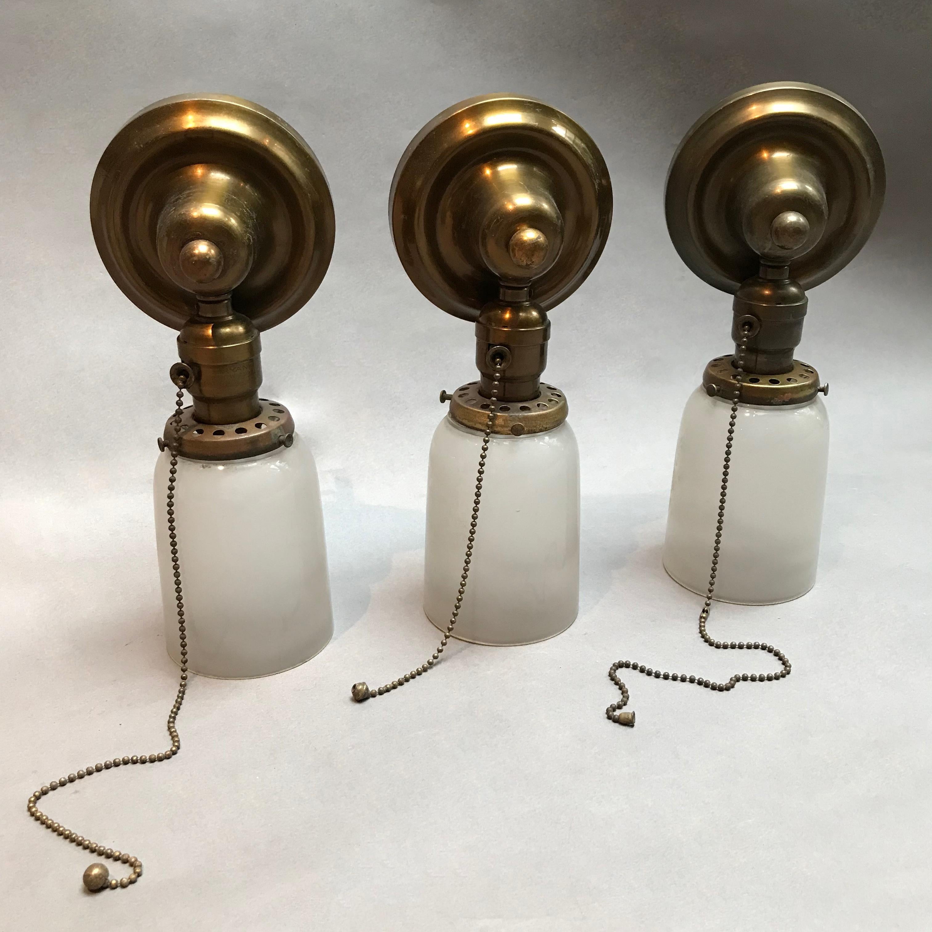 Early 20th century, wall sconce lights feature brass back plates and pull chain fitters with frosted glass bell shades are newly wired to accept up to 100 watt bulbs each. The back plate measures 4.5 inches diameter, the shade itself is 4.5 inches