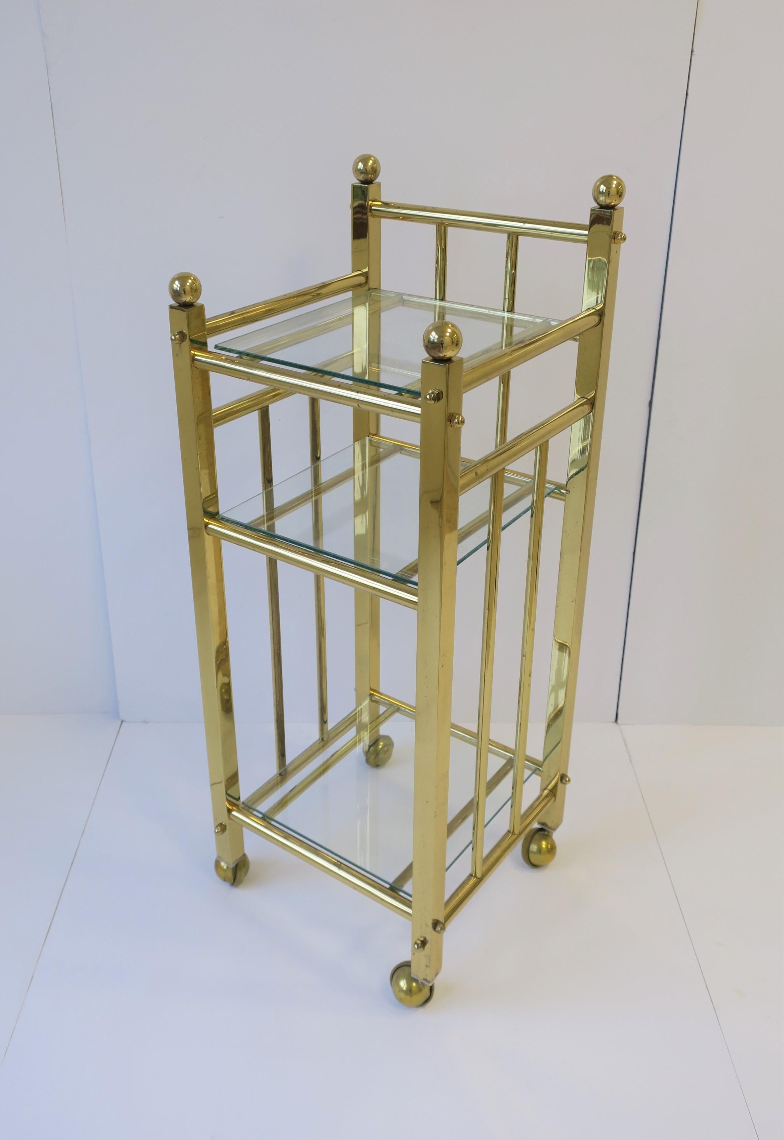A brass and glass bar cart or étagère with shelf on brass caster wheels. Piece can work well as a small bar cart, bathroom caddy, etc. 

Piece measures: 13 in. x 13 in. x 33.5 in. H

    