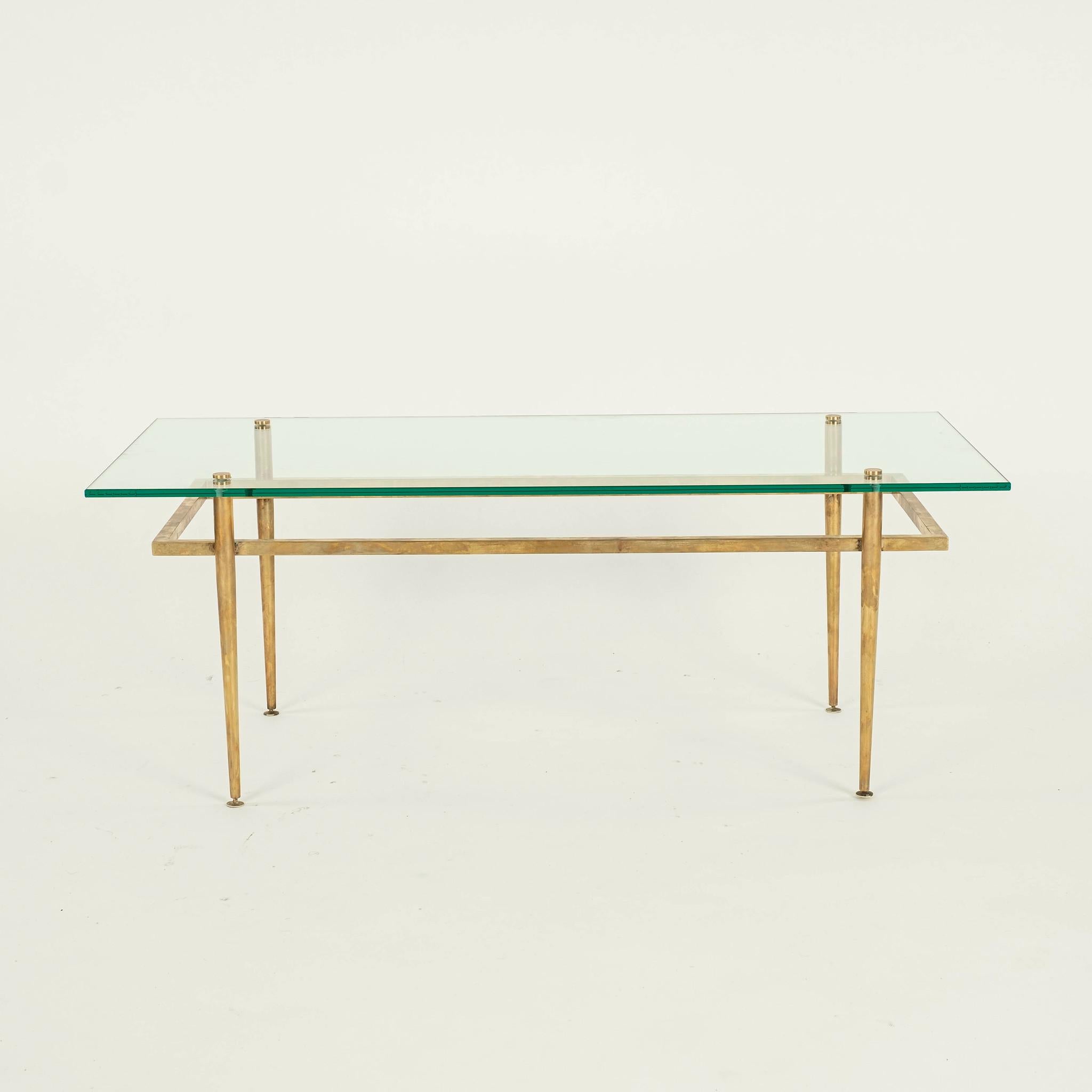Brass and glass cocktail table featuring tapered cylindrical legs with square brass tube bracing, a glass top, and showcasing natural patina.