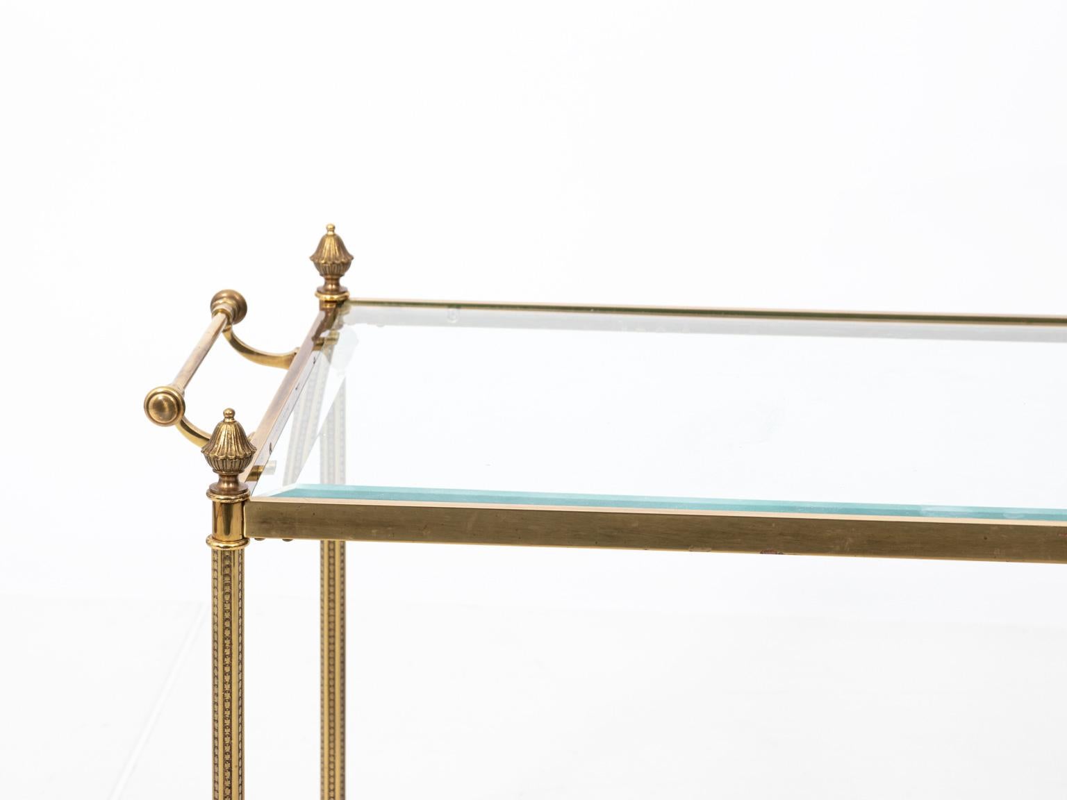 Polished brass and glass coffee table with two handles, circa 1970s. The piece also features beveled glass, pointed finials, and a base cross stretcher. Please note of wear consistent with age including minor oxidation and minor light scratches on