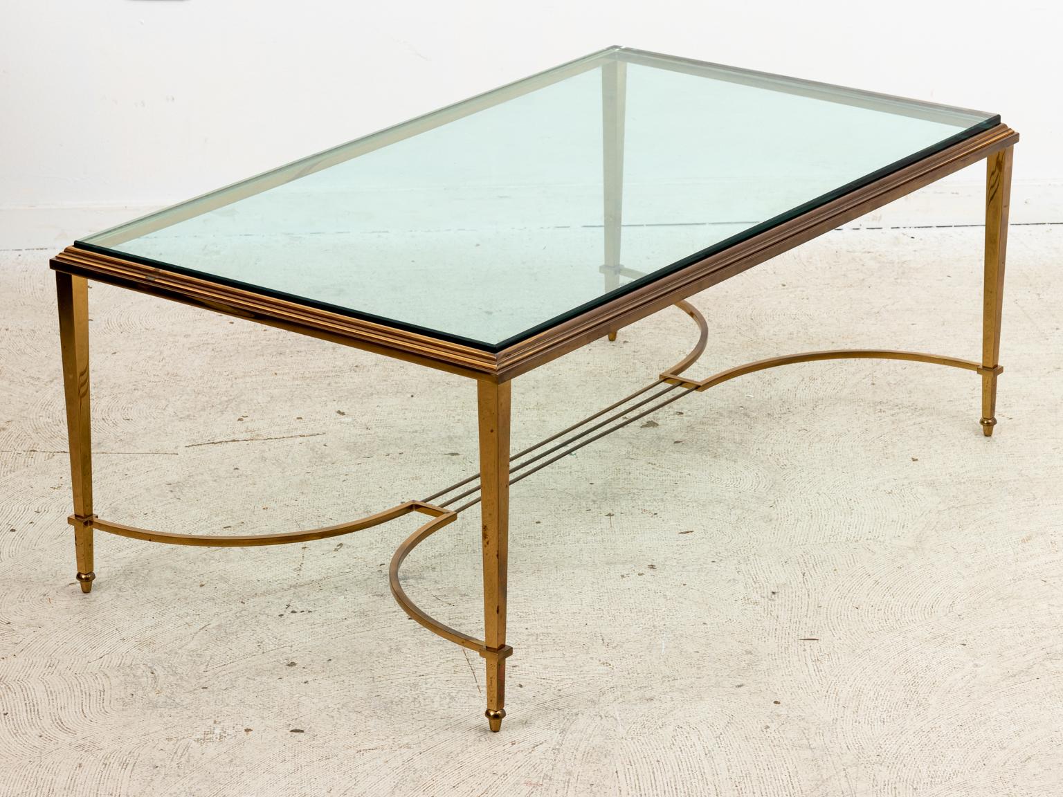 Glass top and brass frame coffee table with bottom c-shaped cross stretcher. The table is accented with molded trim on the tabletop and arrow feet. Please note of wear consistent with age including patina and oxidation to the metal.