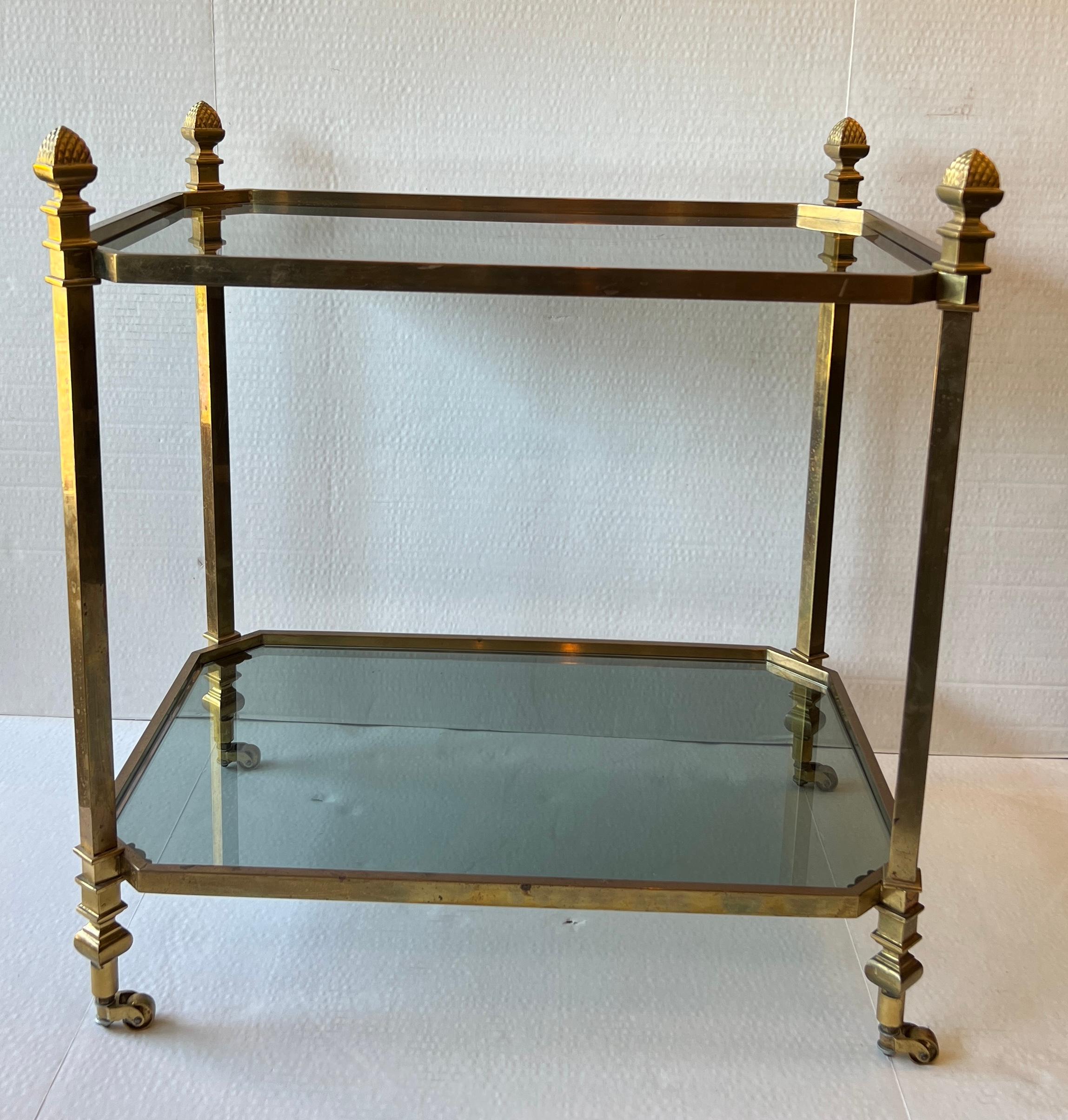 Beautiful gold tone brass table with four large pinecone finials on casters. Two shelves of grey toned glass

