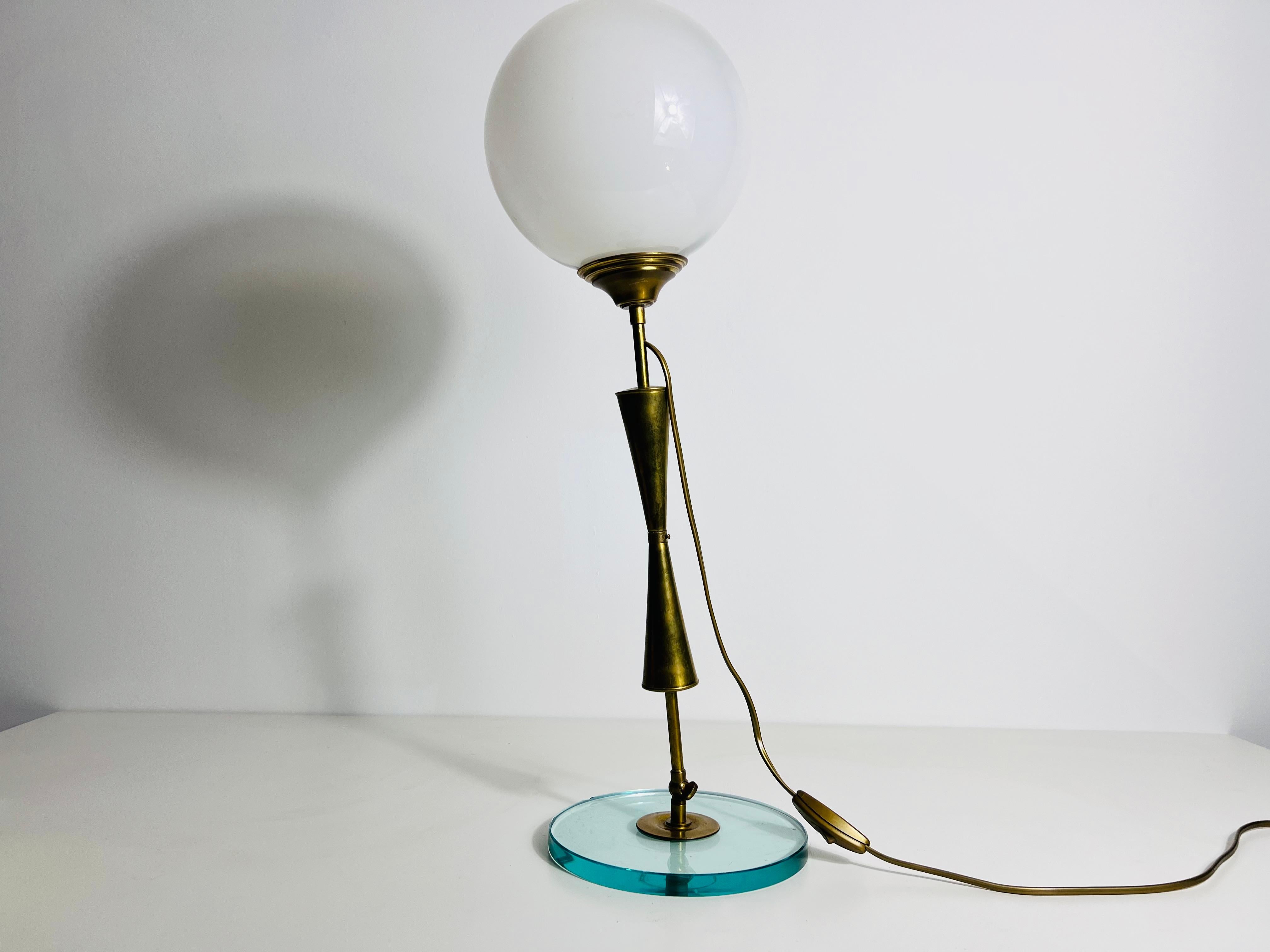 Questa squisita lampada da tavolo degli anni '60 in Italia è un vero capolavoro della metà del secolo scorso, che combina vetro trasparente, ottone e vetro opalino in un design sorprendente. La lampada è caratterizzata da una base rotonda in vetro