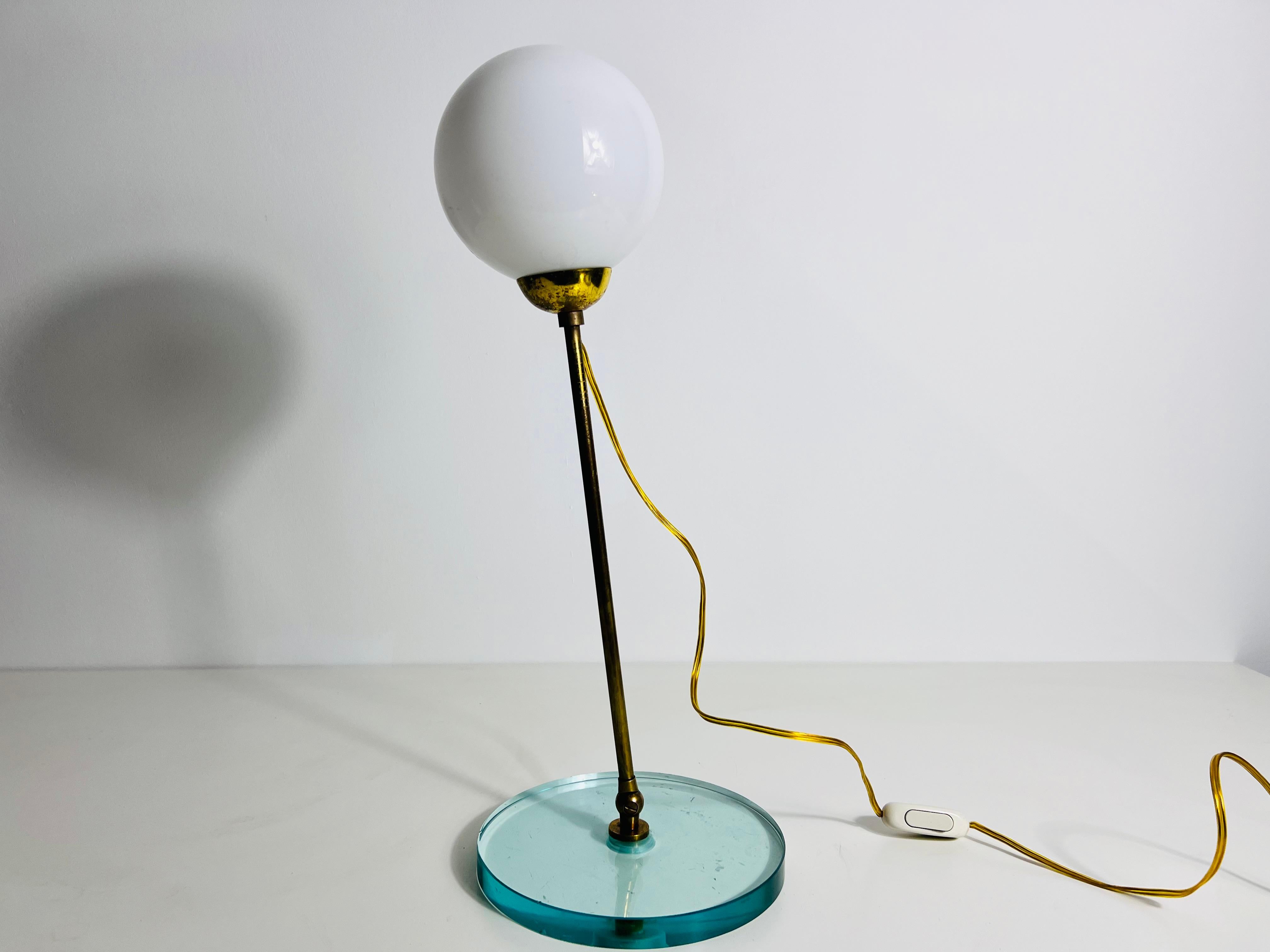 Questa squisita lampada da tavolo degli anni '60 in Italia è un vero capolavoro della metà del secolo scorso, che combina vetro trasparente, ottone e vetro opalino in un design sorprendente. La lampada è caratterizzata da una base rotonda in vetro