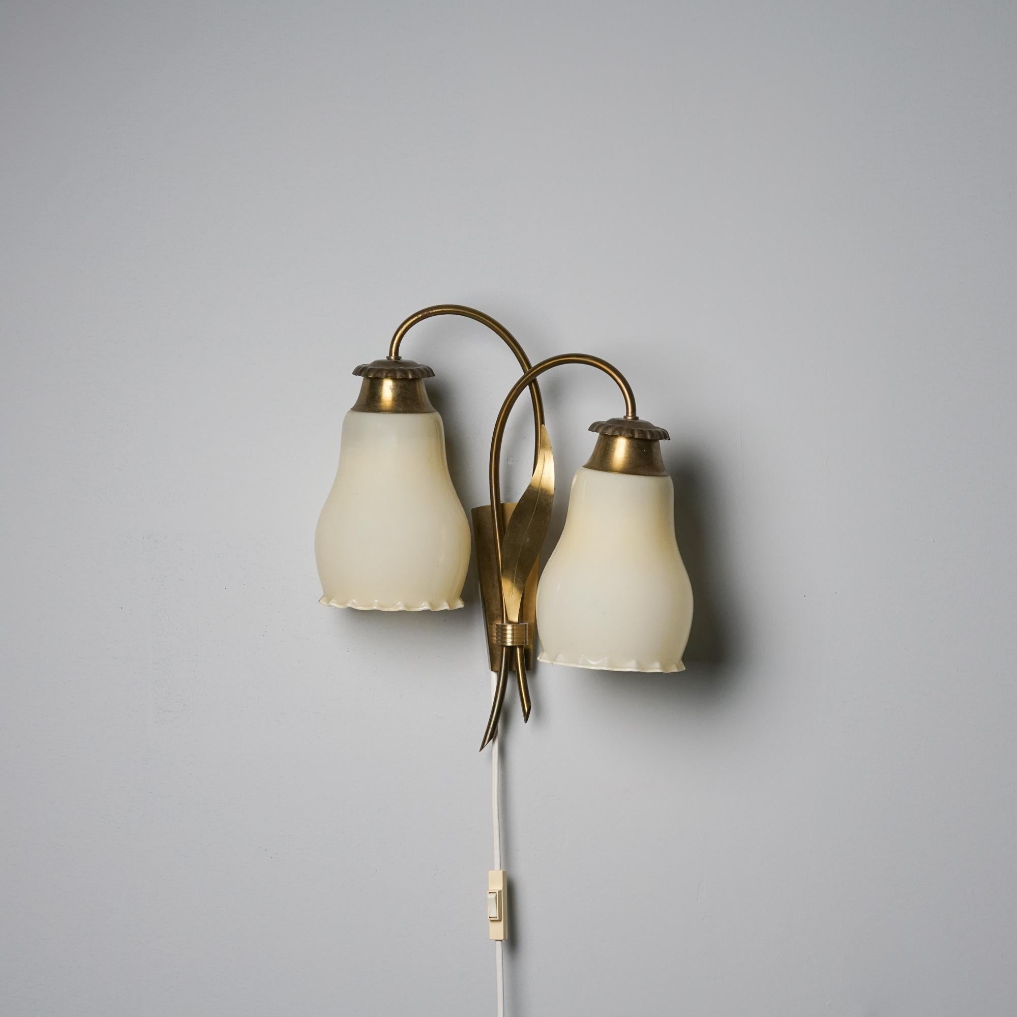 Lampada da parete a due bracci, prodotta in Finlandia, anni '50. Paralumi in vetro opalino con struttura in ottone. Buone condizioni d'epoca, lieve patina coerente con l'età e l'uso. 