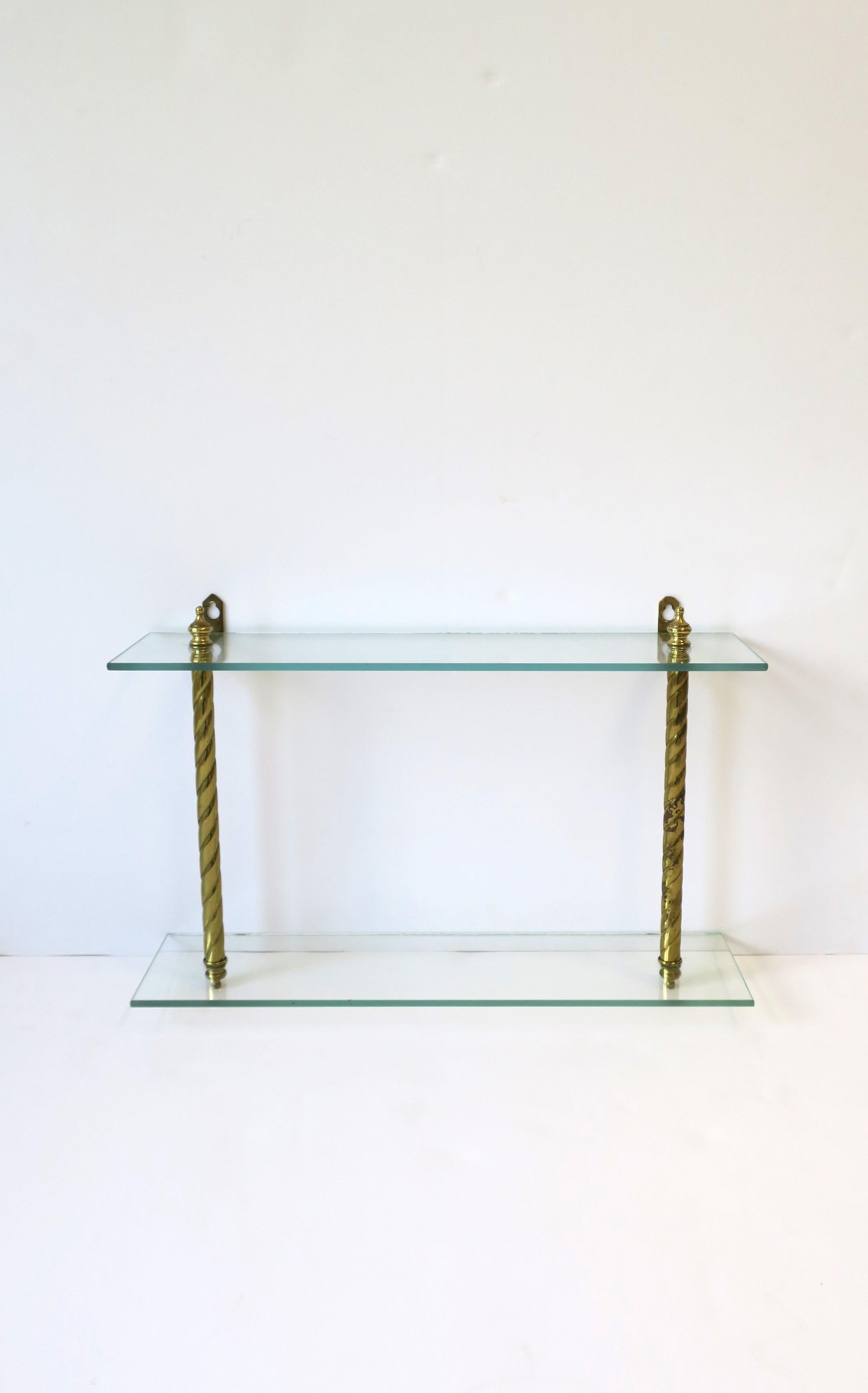 A brass and glass wall shelf with finial design, circa mid-20th century. Piece has two glass shelves and brass hardware with finial detail at top and bottom, including brass hook hardware at back for hanging. Piece may hold items in a bathroom,