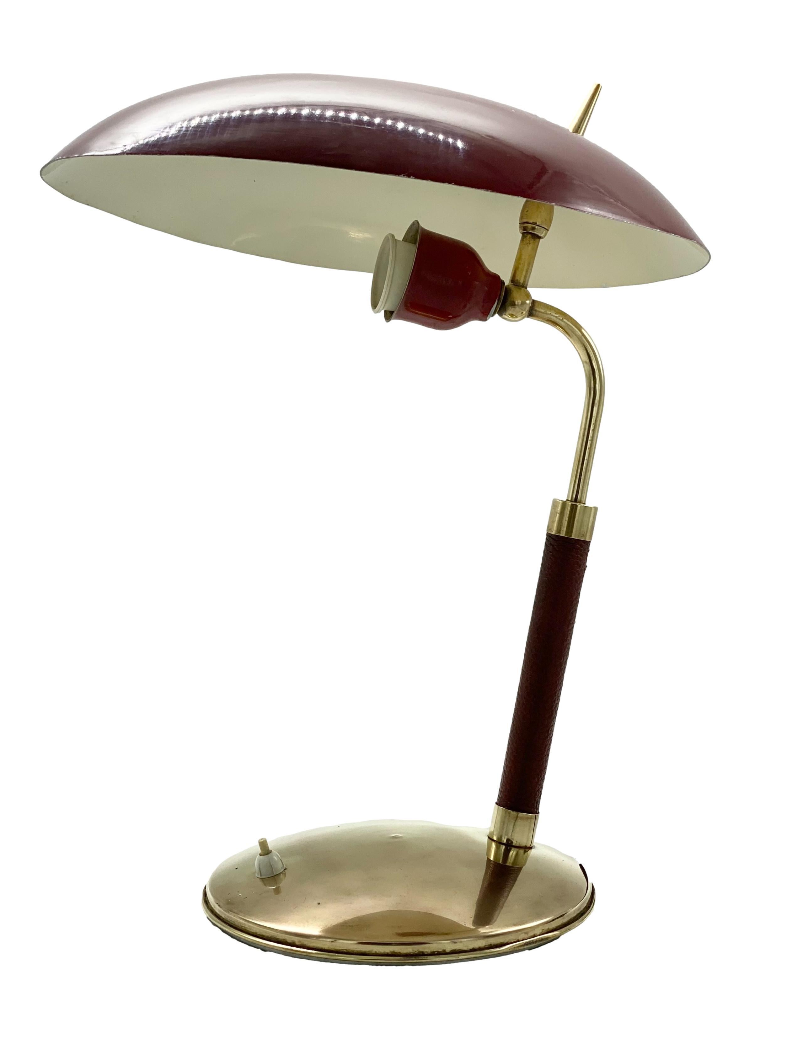 Table lamp, red metal, brass and leather, Lumen Italia, 1950s. 
This colourful desk lamp is of balanced, organic design and elegant appearance. 
On the round brass base there is a switch. The lamp shows an overall patina which contributes to the